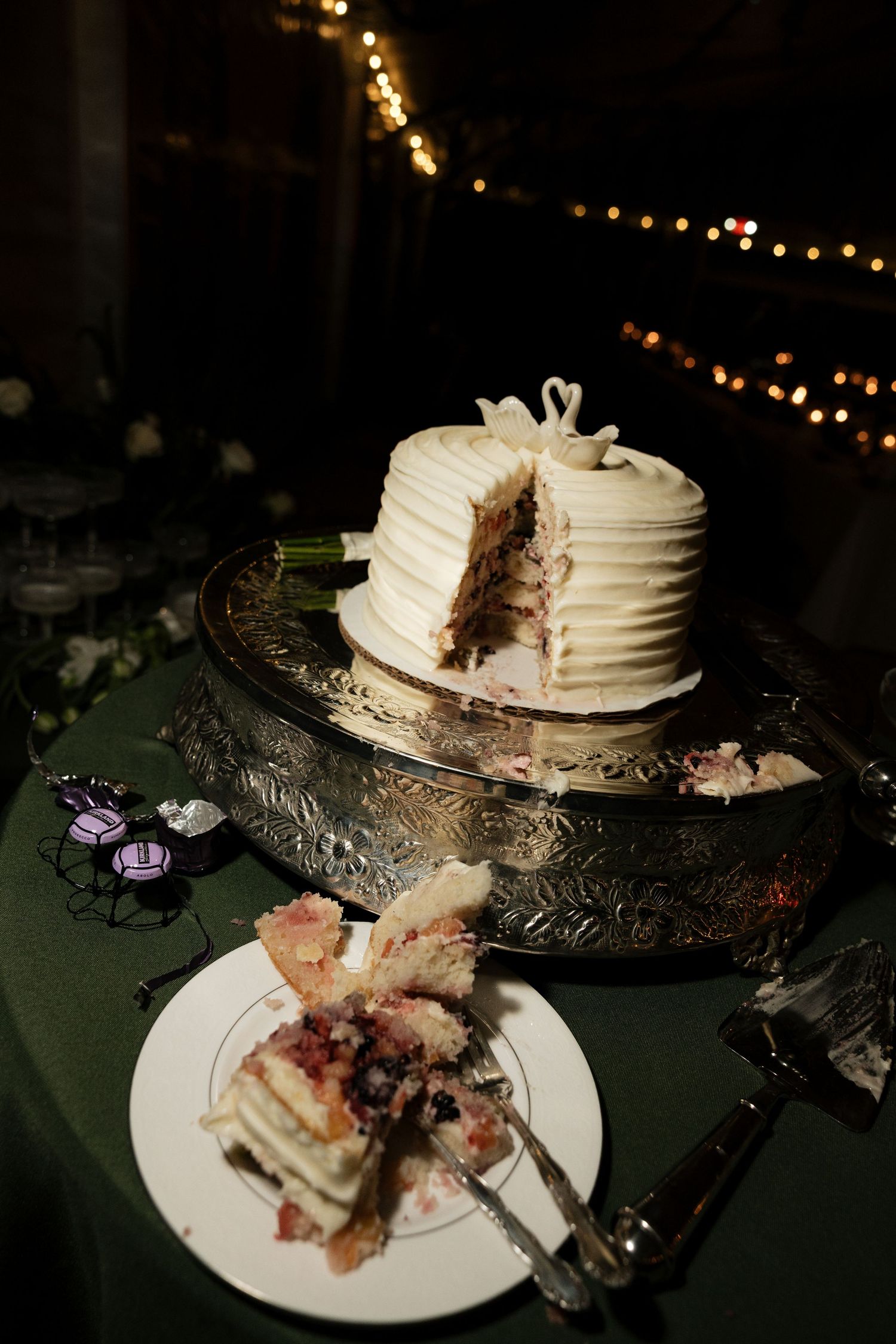 Partially sliced white wedding cake with textured frosting on a silver ornate cake stand on a green tablecloth.