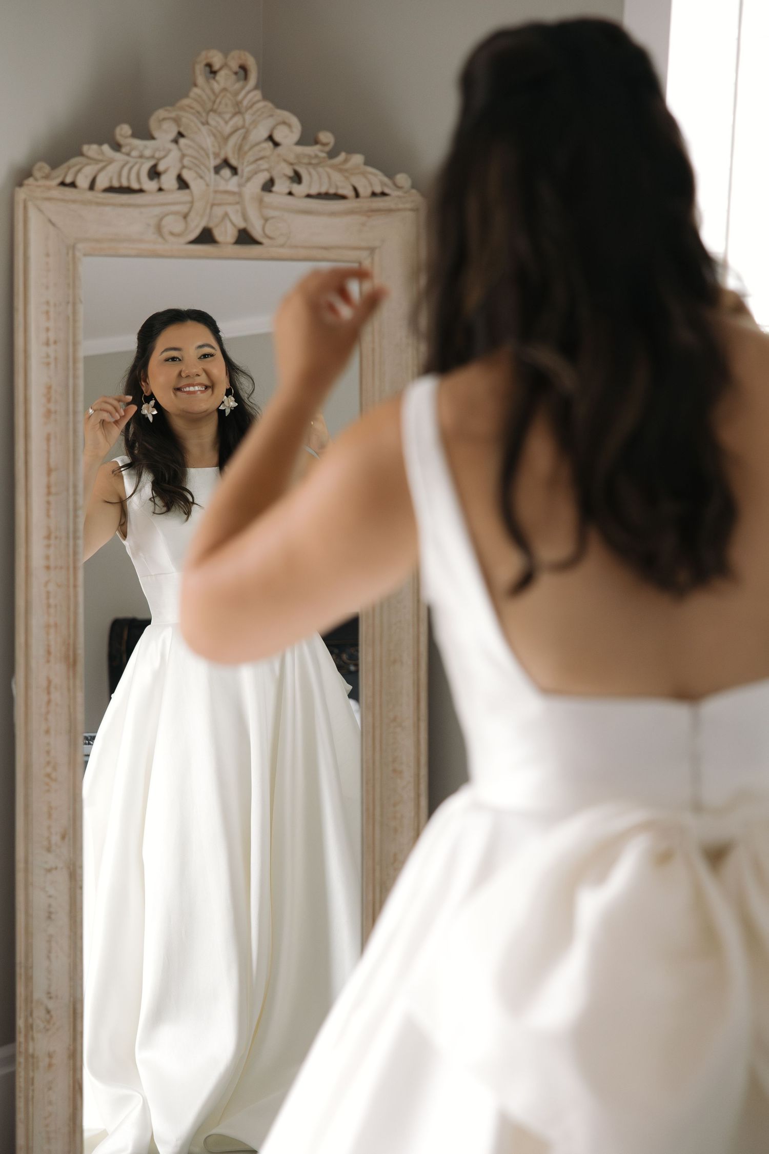 Radiant bride in white satin gown smiling at her reflection in an ornate floor mirror.