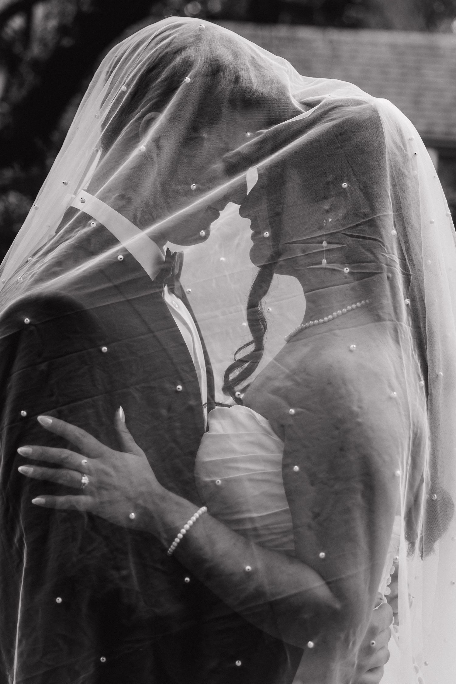Dramatic black and white silhouette of bride and groom embracing beneath flowing pearl-studded wedding veil.