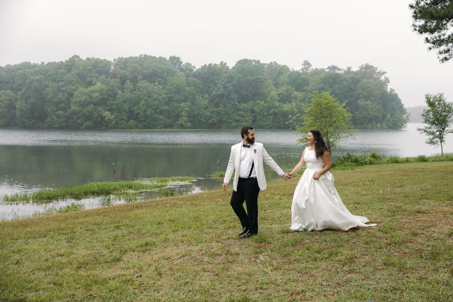 Bride and groom hold hands walking by a misty lake surrounded by lush green trees on their wedding day.