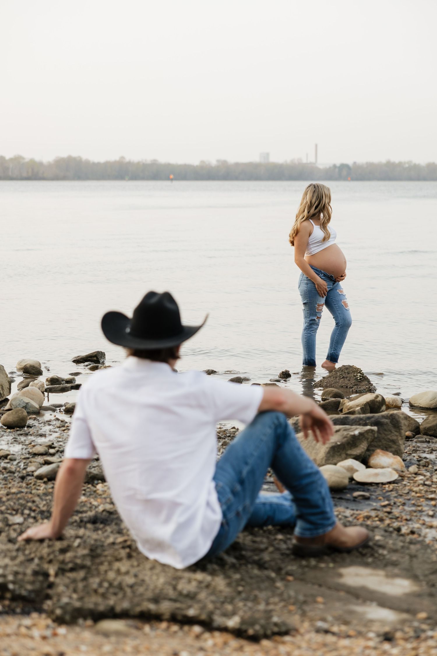 Man in black cowboy hat sits on rocky shore watching pregnant woman stand near river's edge.