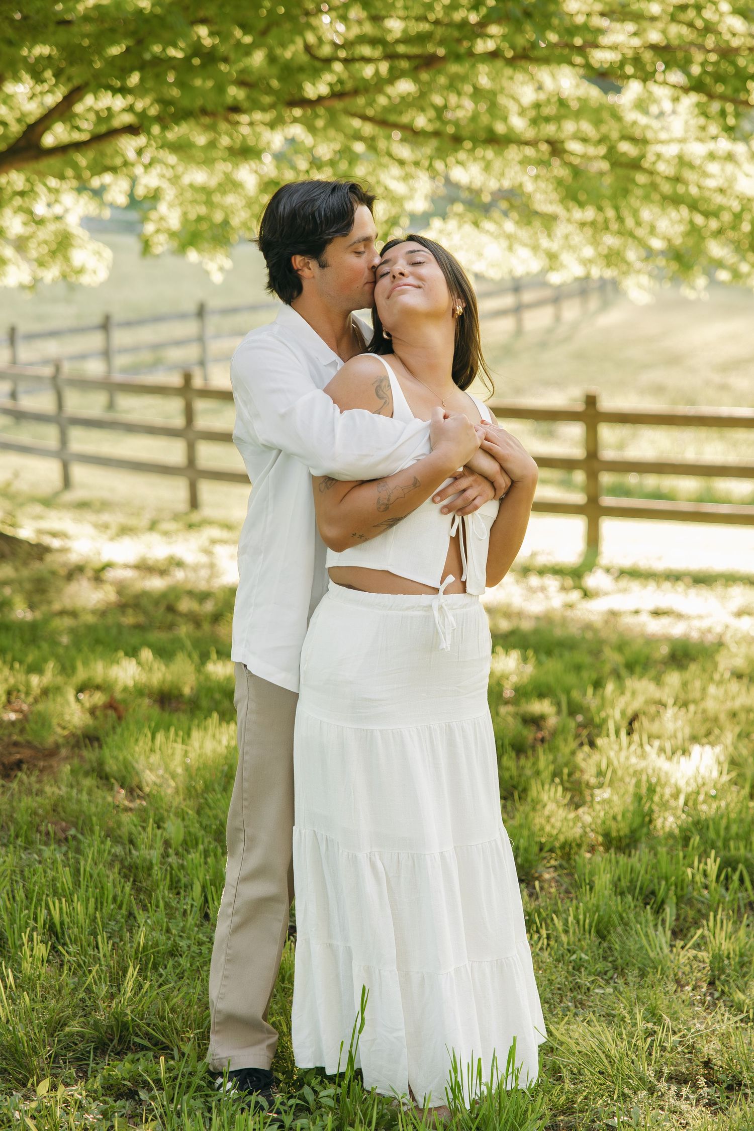Couple embracing outdoors, man kissing woman's cheek as she smiles with eyes closed, both in white near wooden fence.