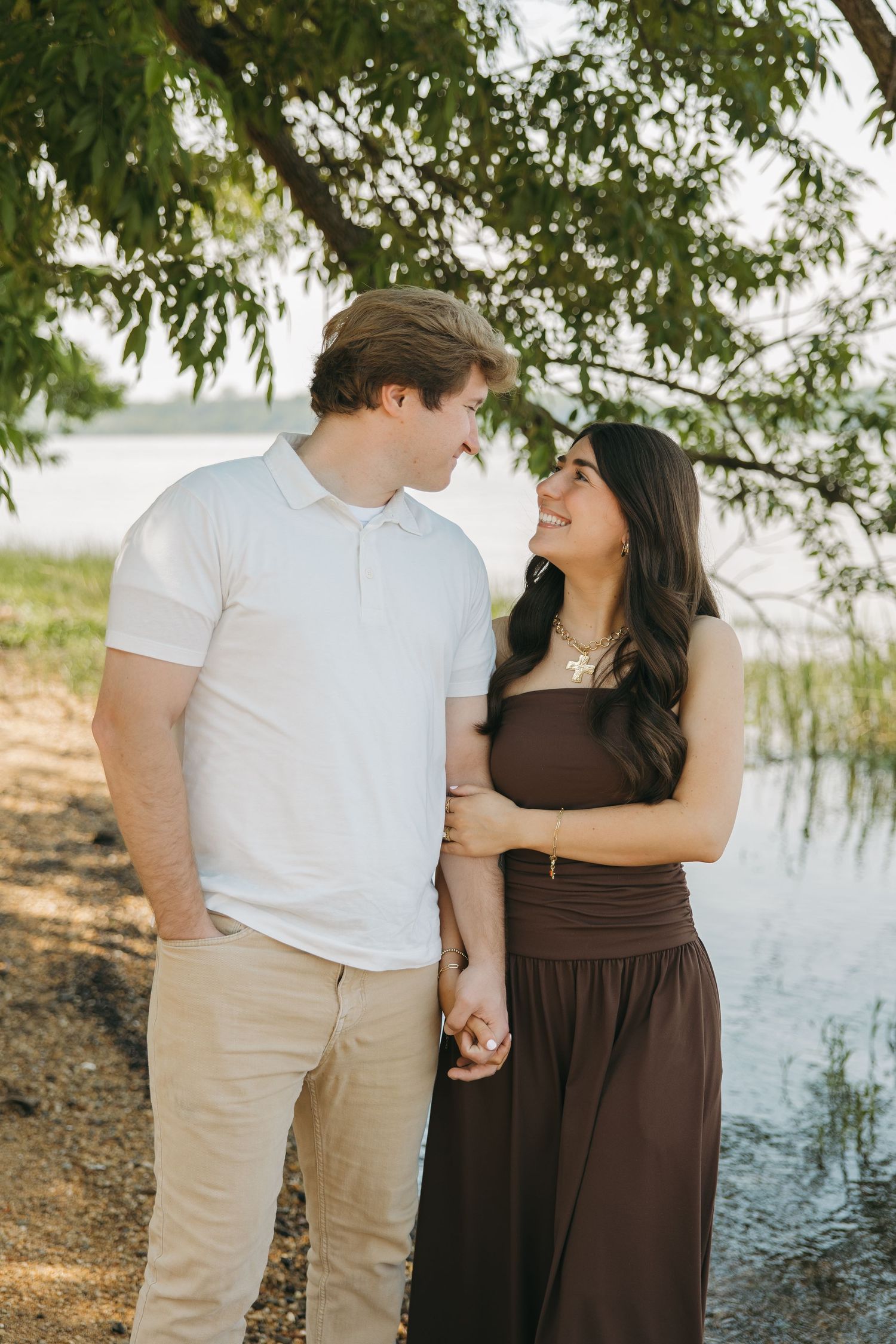 A happy couple gazes at each other lovingly while holding hands near a tranquil lakeside under a tree.