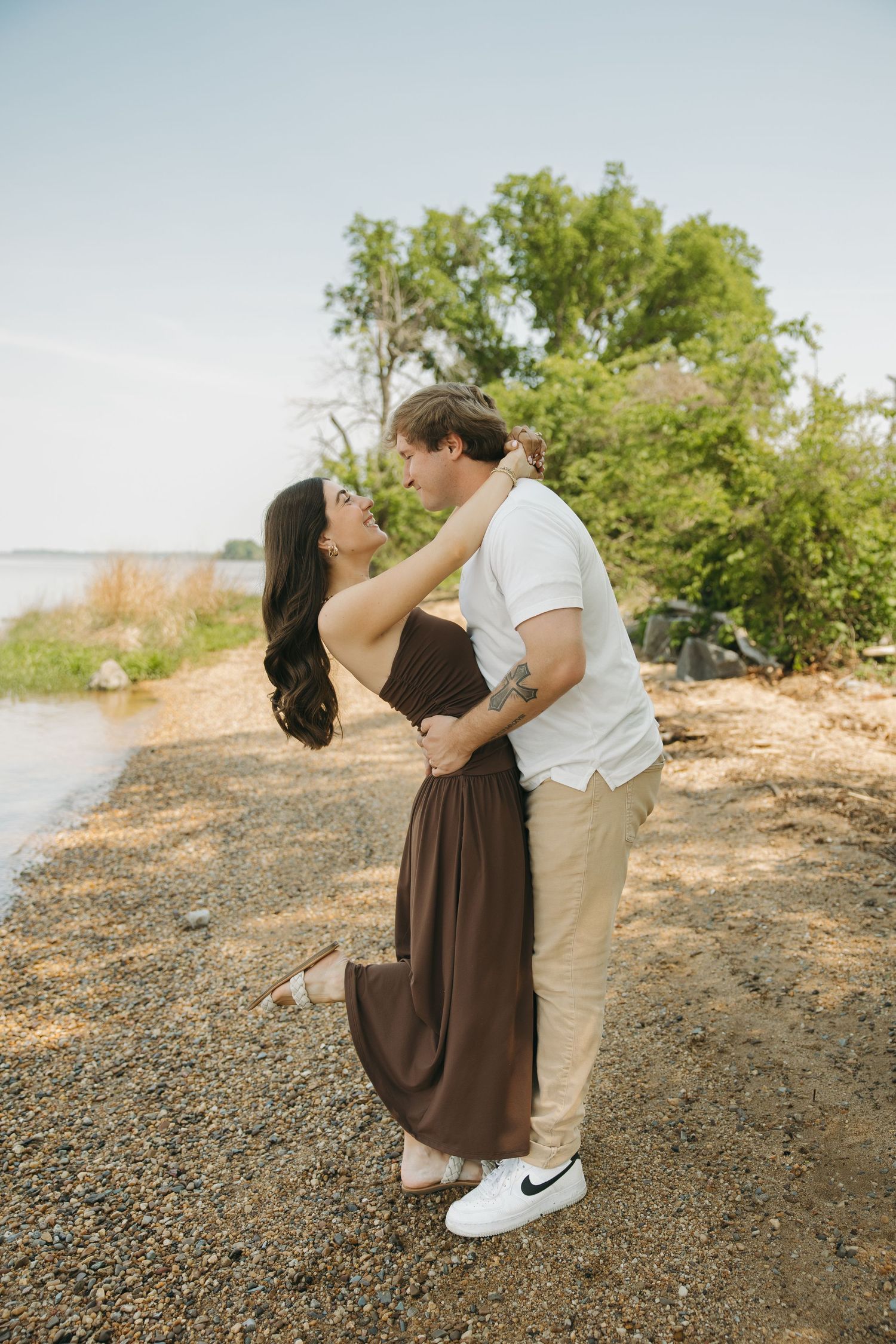 Couple sharing a romantic moment on a rocky shoreline with lush green trees behind.