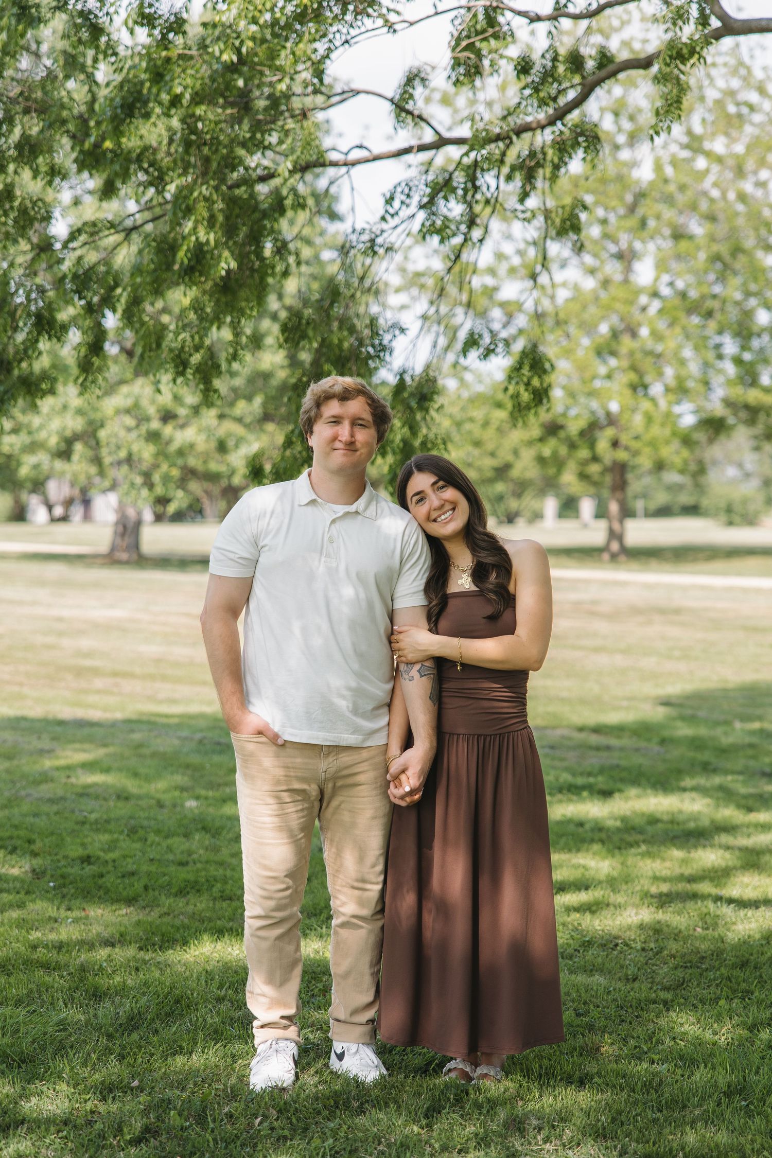 Smiling couple standing together holding hands under a tree in a green park.