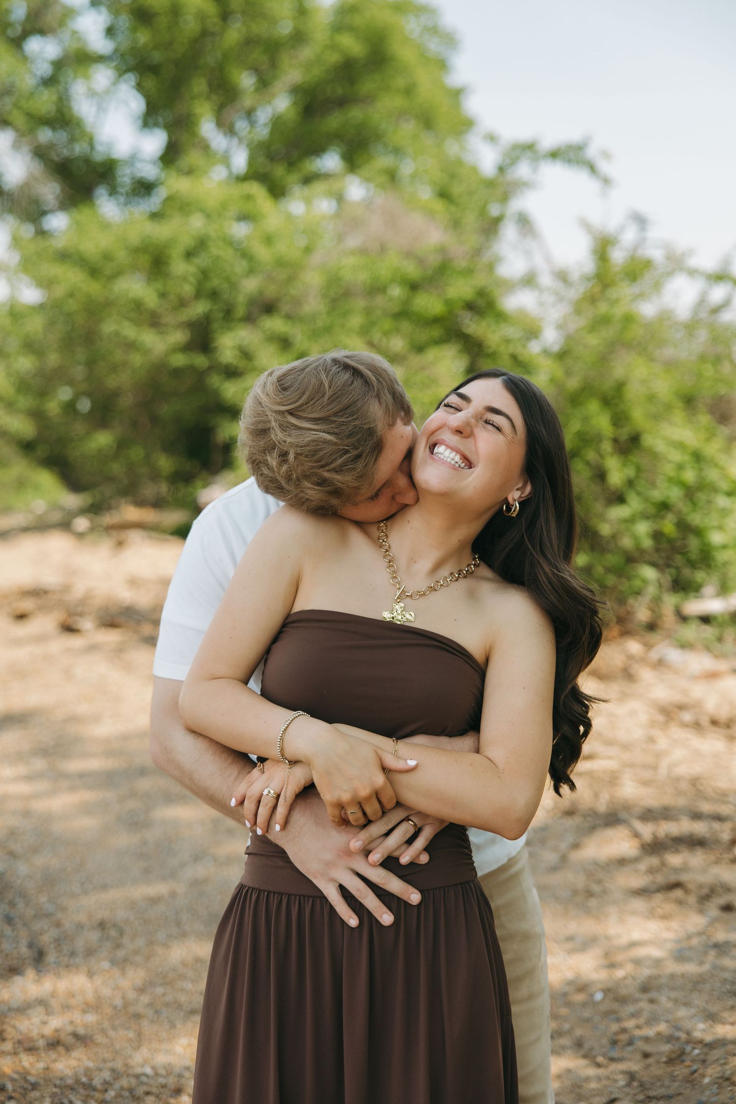 A couple embraces outdoors, woman in brown strapless dress laughing joyfully as man kisses her cheek.