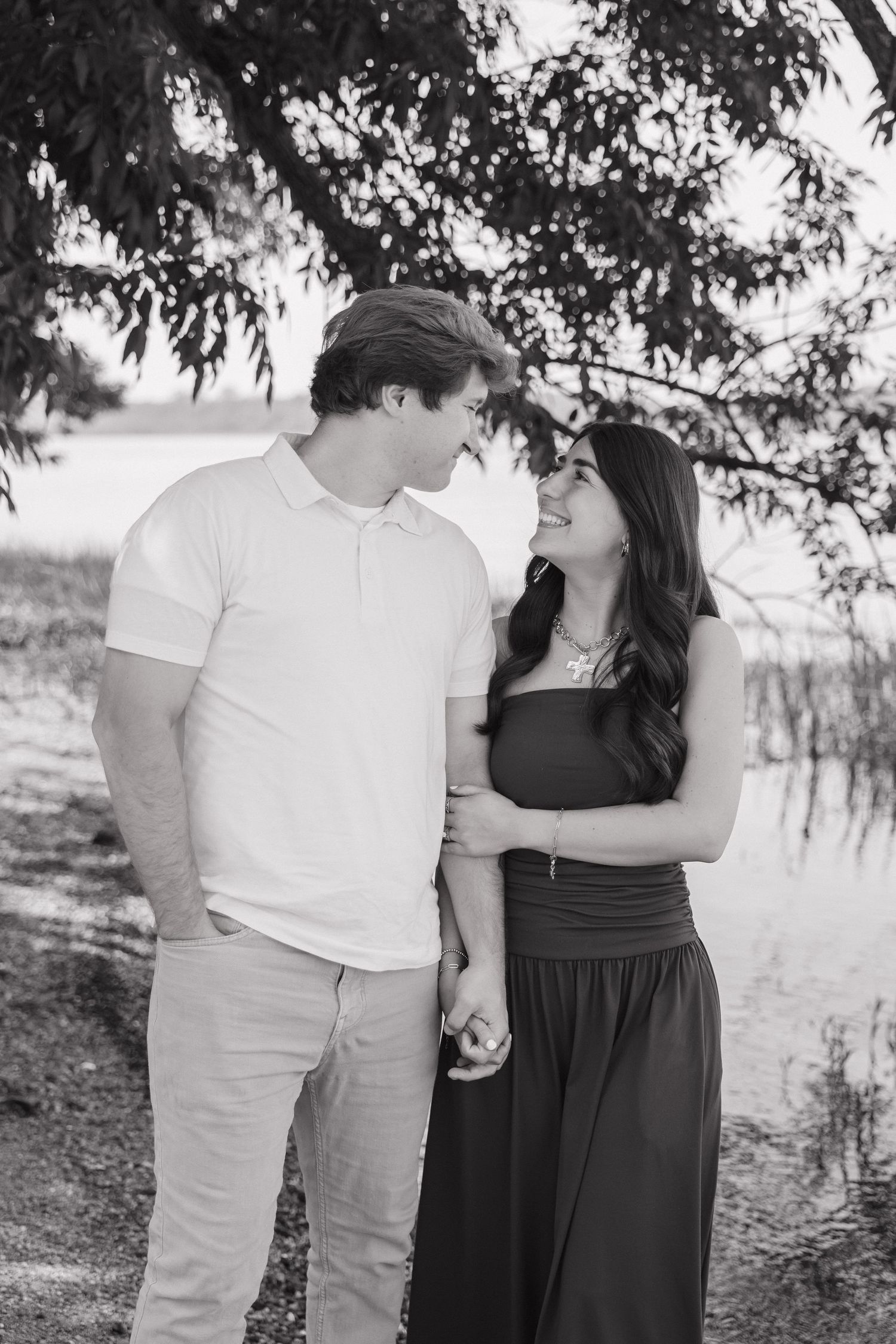 A black and white photo of a couple smiling at each other while holding hands near a peaceful lakeside.