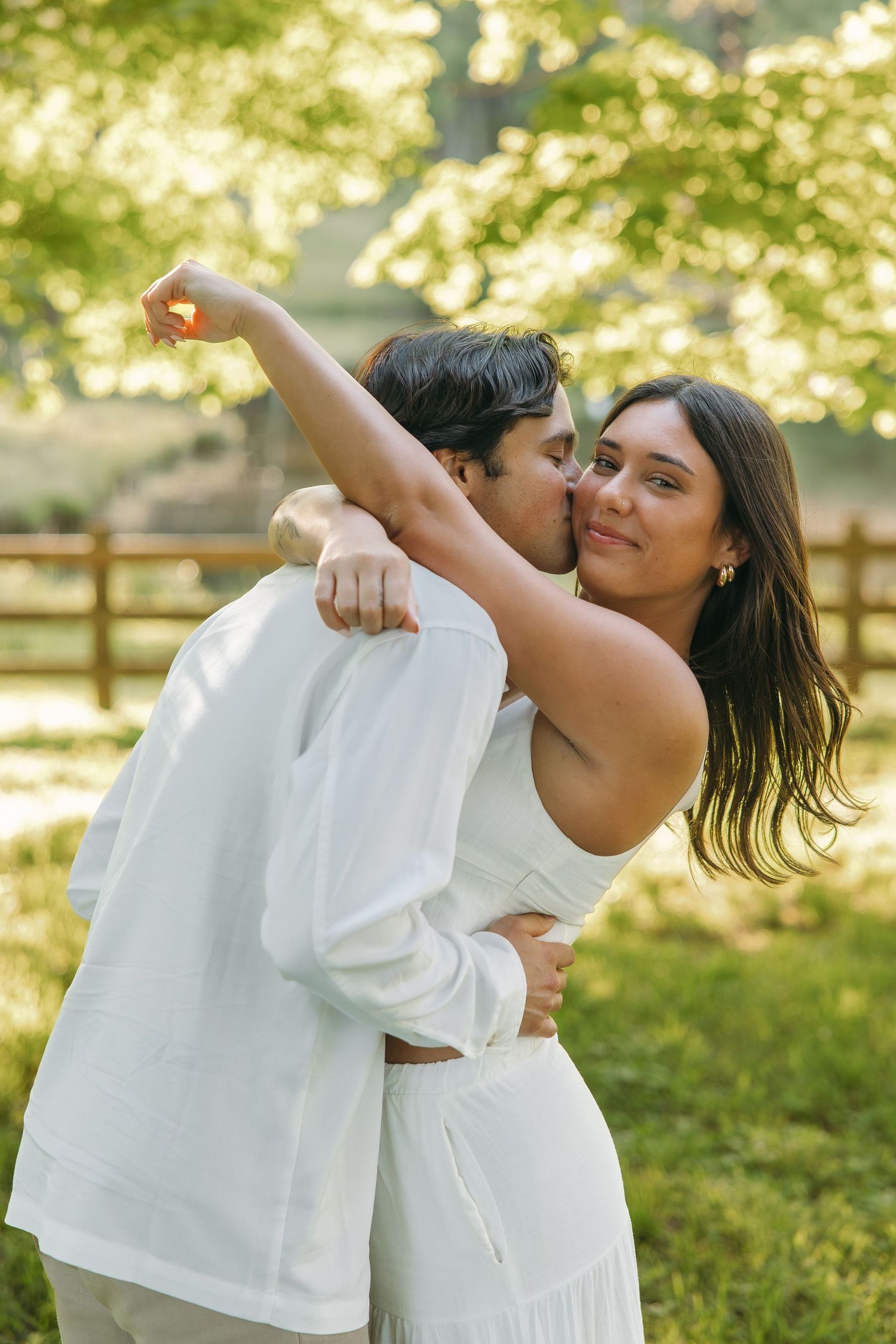 Romantic couple dancing outdoors, woman raising arm while man kisses her cheek, surrounded by lush green trees.