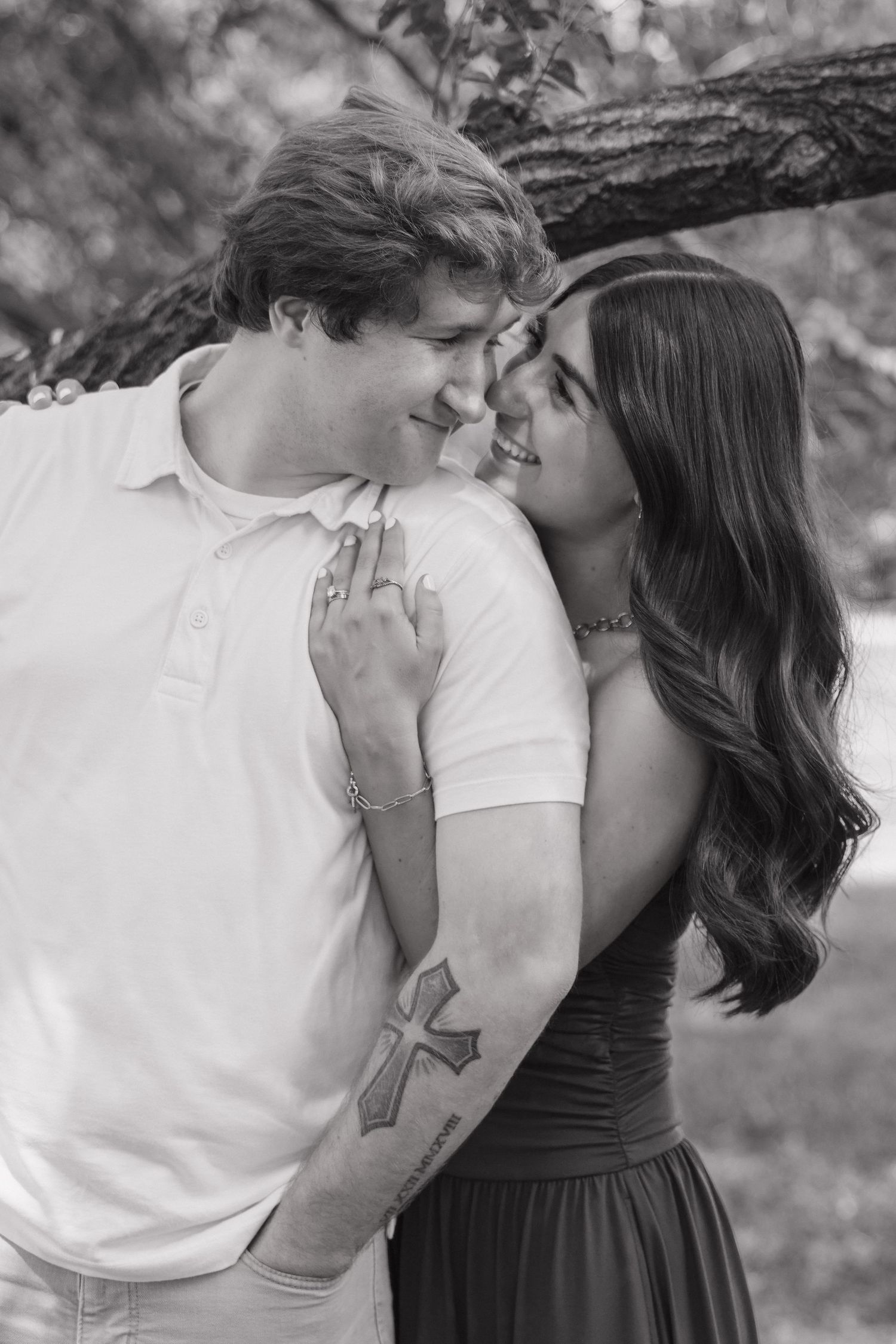 Black and white photo of couple smiling at each other outdoors, man has cross tattoo on arm.