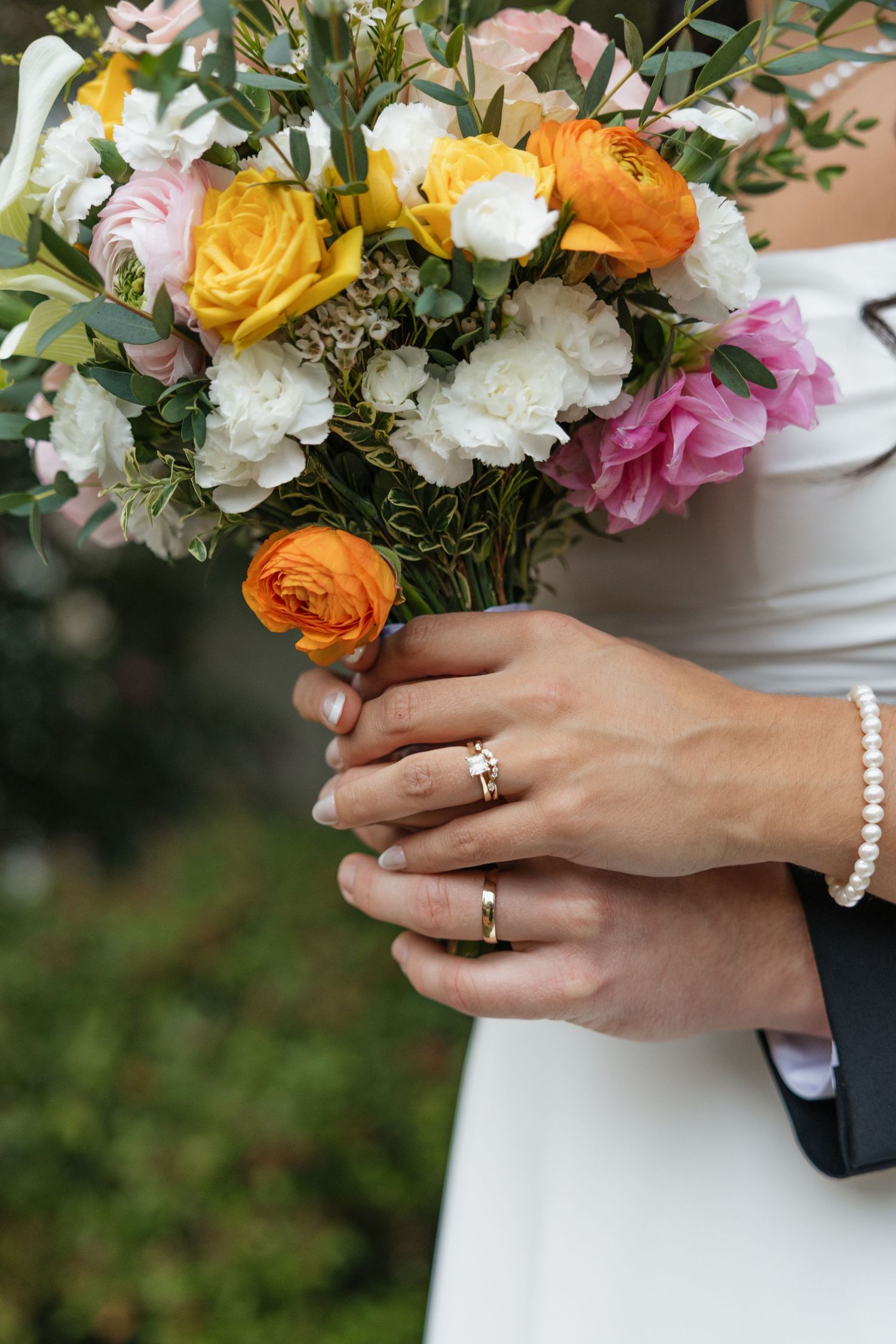 Bride and groom holding a colorful wedding bouquet with orange, pink, and white roses, showing their gold wedding rings.