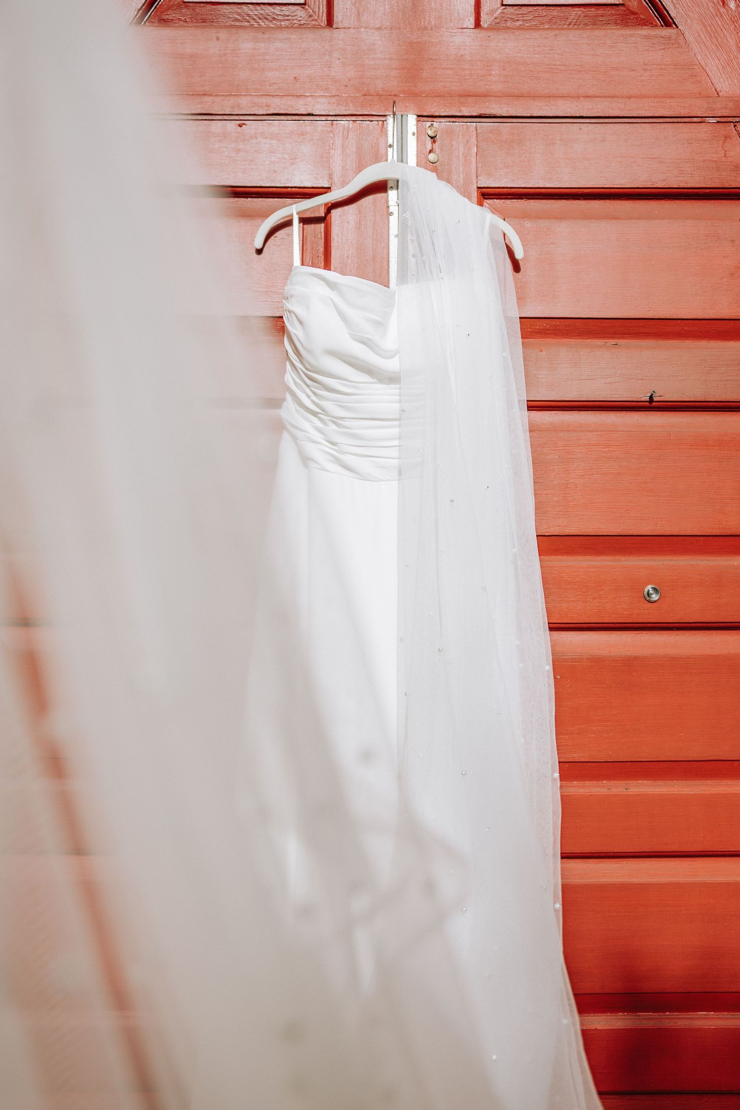 White strapless wedding gown with veil hanging on hanger against red wooden door.