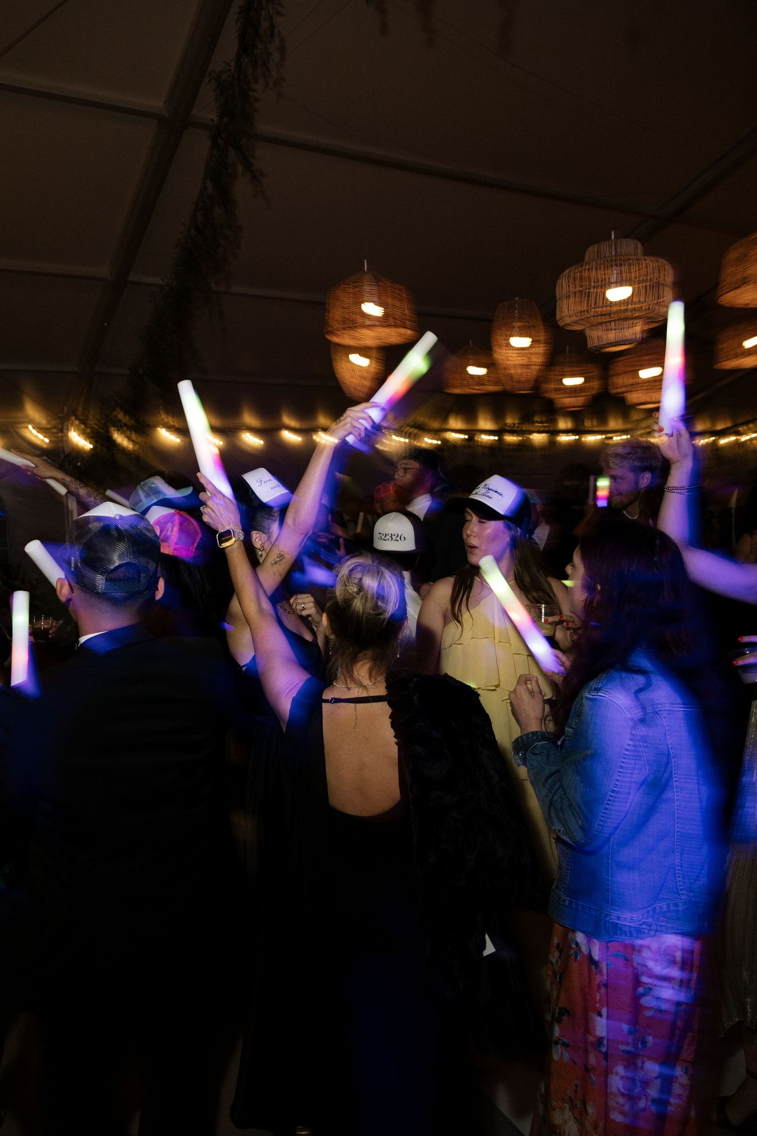 Wedding guests dancing and waving glowing LED foam sticks under wicker lights at night reception.