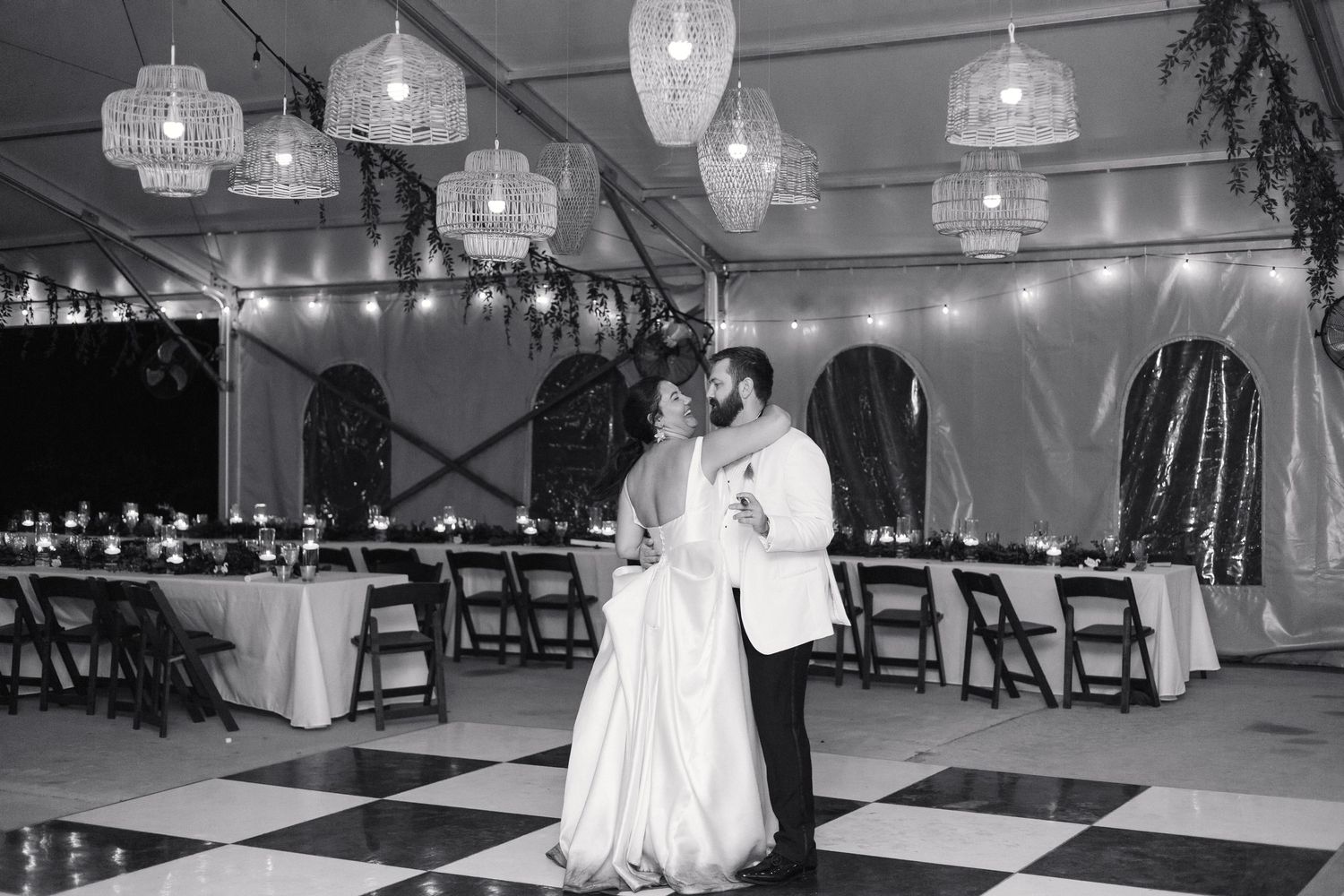 Bride and groom share first dance on checkered floor under chandelier lights in elegant black and white wedding reception tent.
