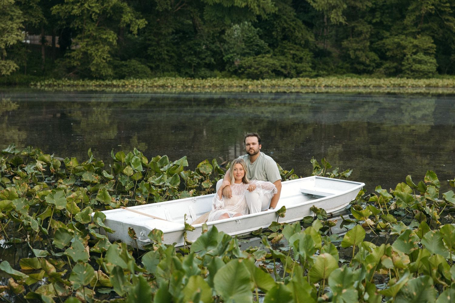Happy couple relaxing in white rowboat amid lush lily pads on tranquil lake with dense green forest backdrop.