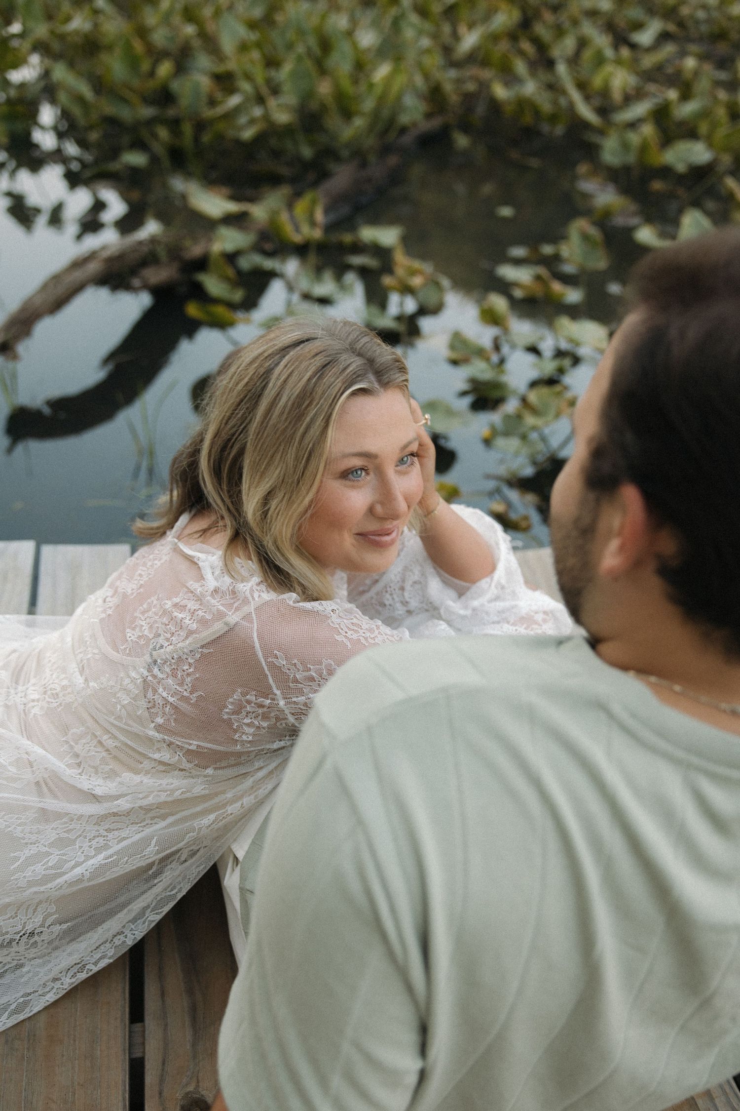Woman in white lace dress leaning on dock railing smiling at man, with water and lily pads in background.