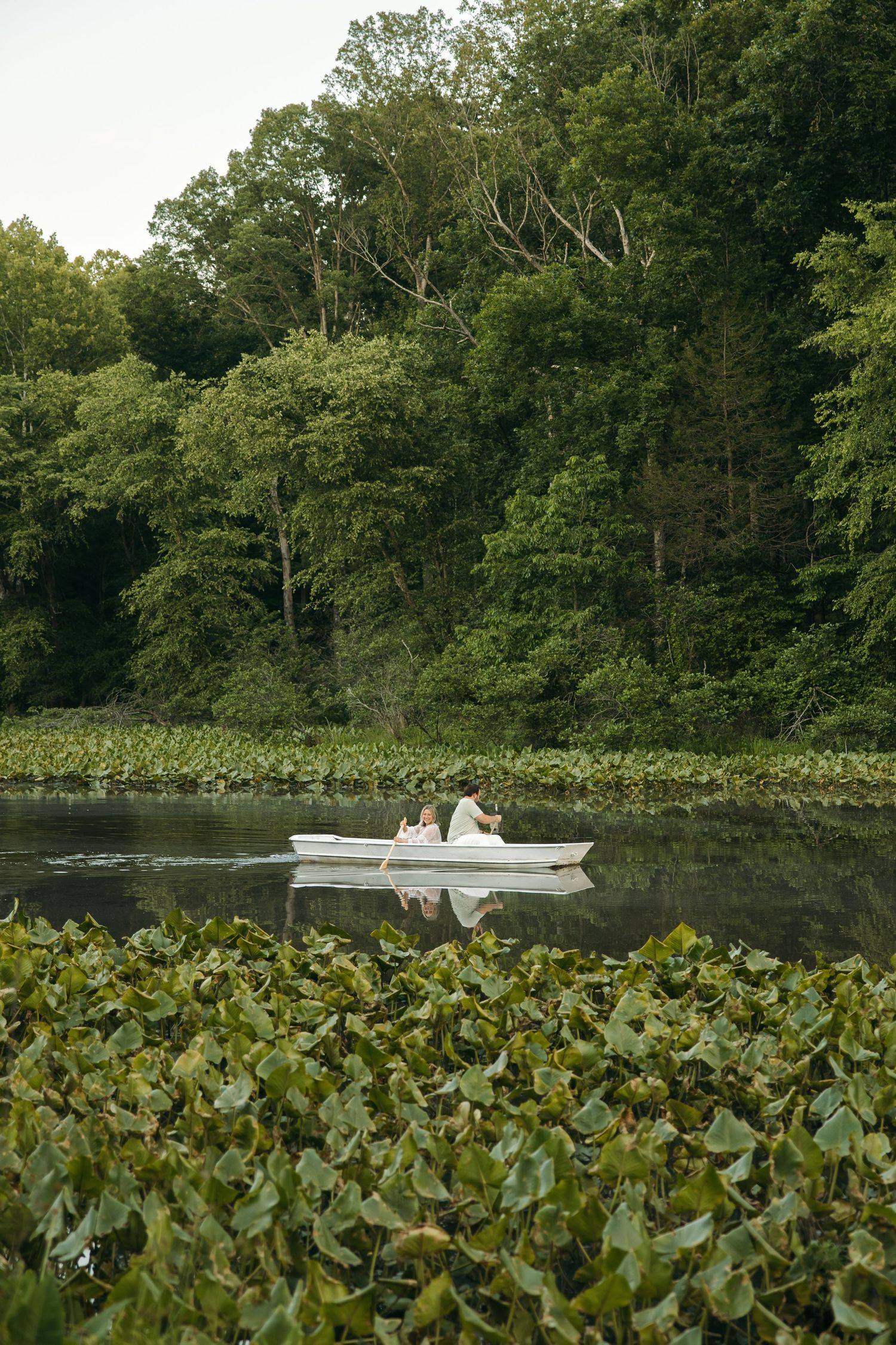 Couple rowing a white boat on a tranquil lake surrounded by lily pads and dense green forest trees.