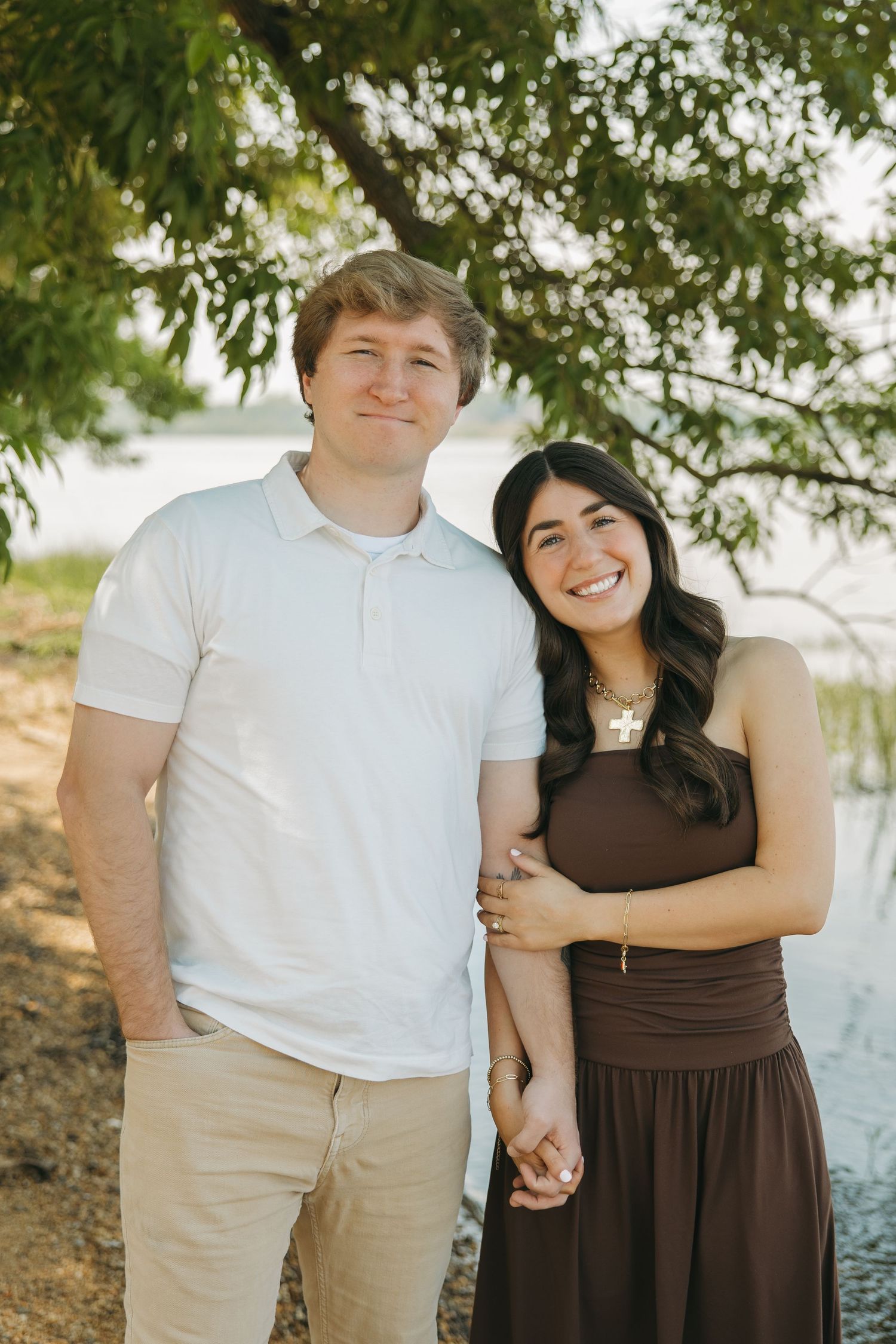 A smiling couple holds hands under a shady tree near a sunlit lake during an outdoor photo session.