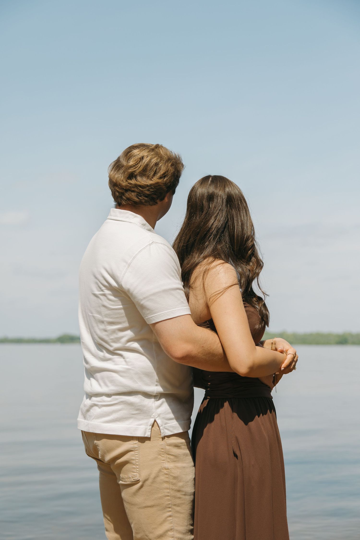 Couple standing together from behind, holding hands while overlooking a calm lake.