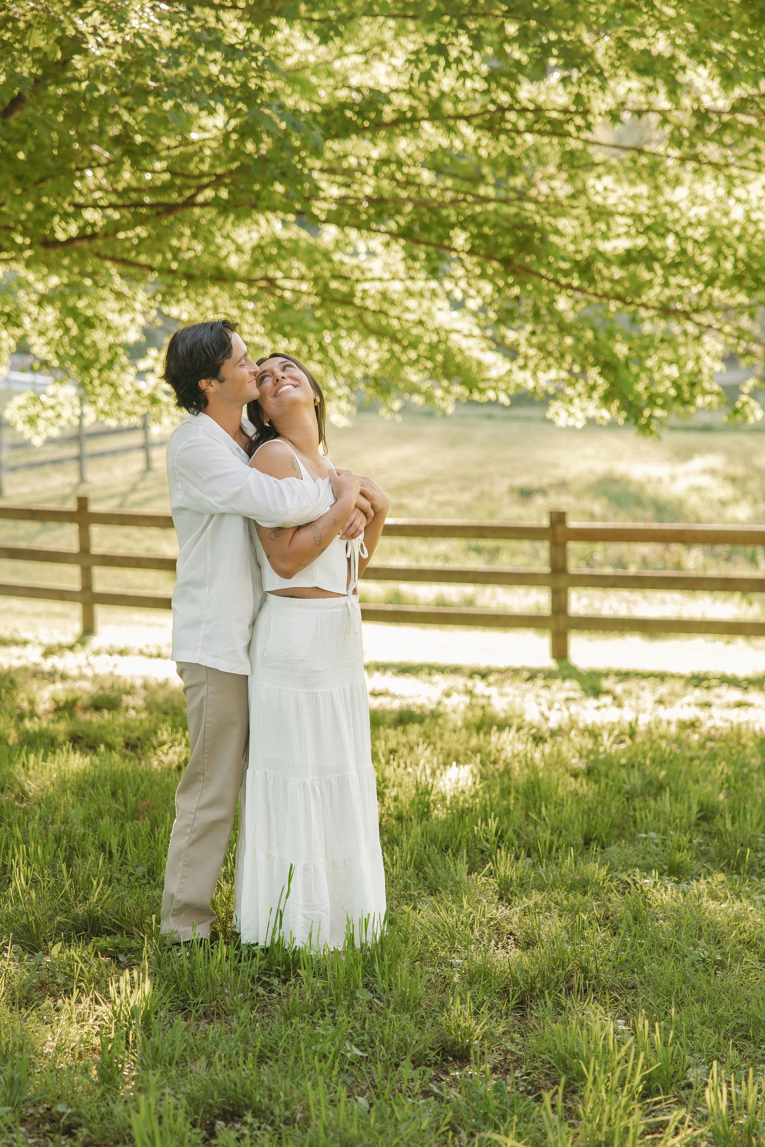 Happy couple embracing and laughing under a large green tree in a sunny pastoral setting.