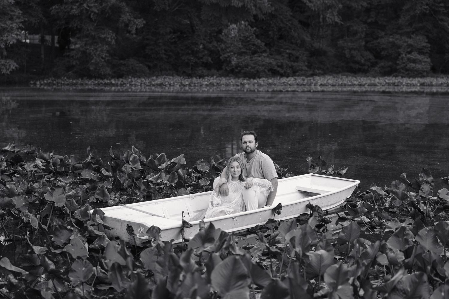 Black and white photo of a couple sitting closely together in a white rowboat on a lily pad-covered lake.