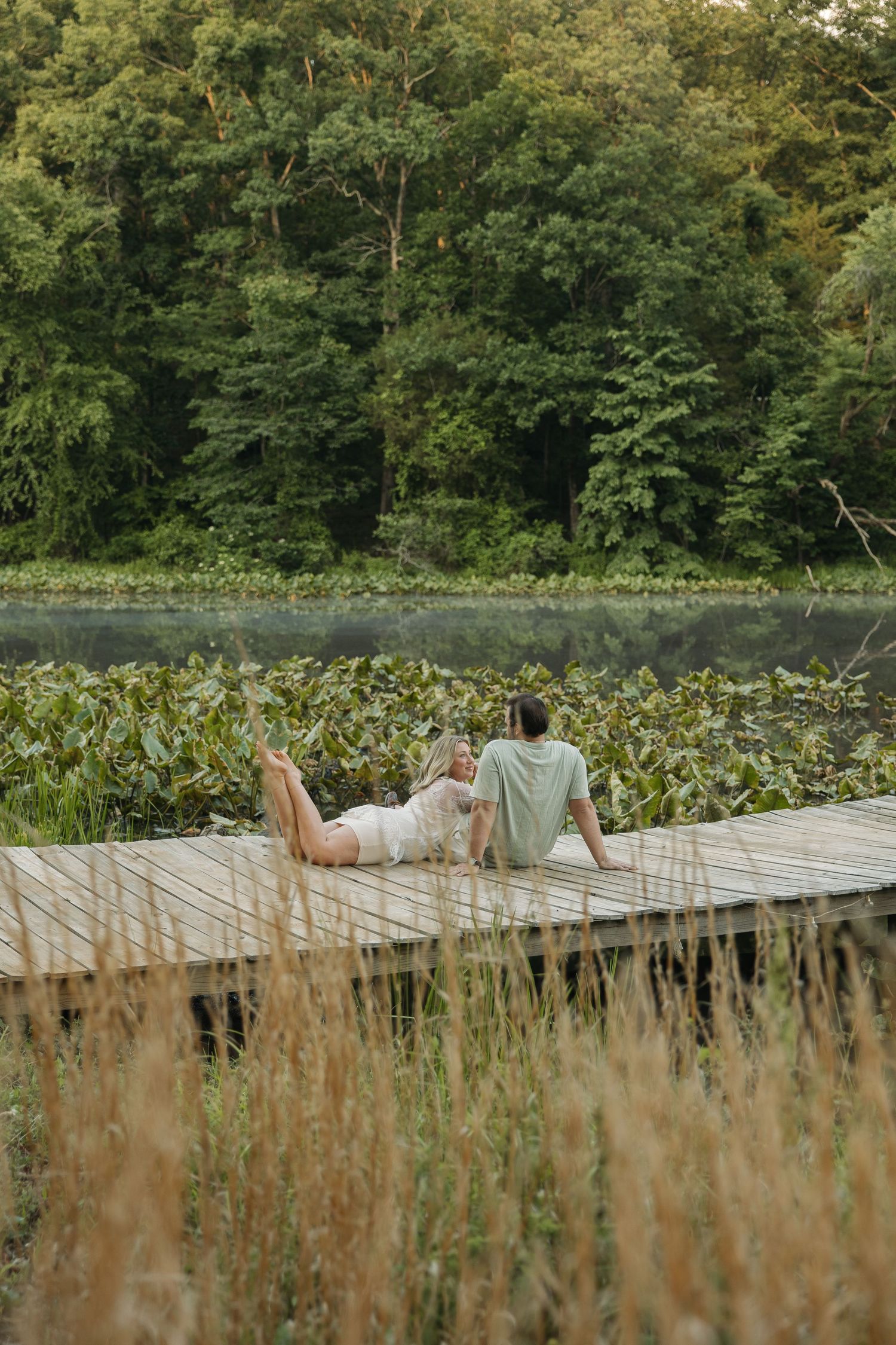 Couple lying and sitting together on a wooden lakeside dock surrounded by lush green trees and tall grass.