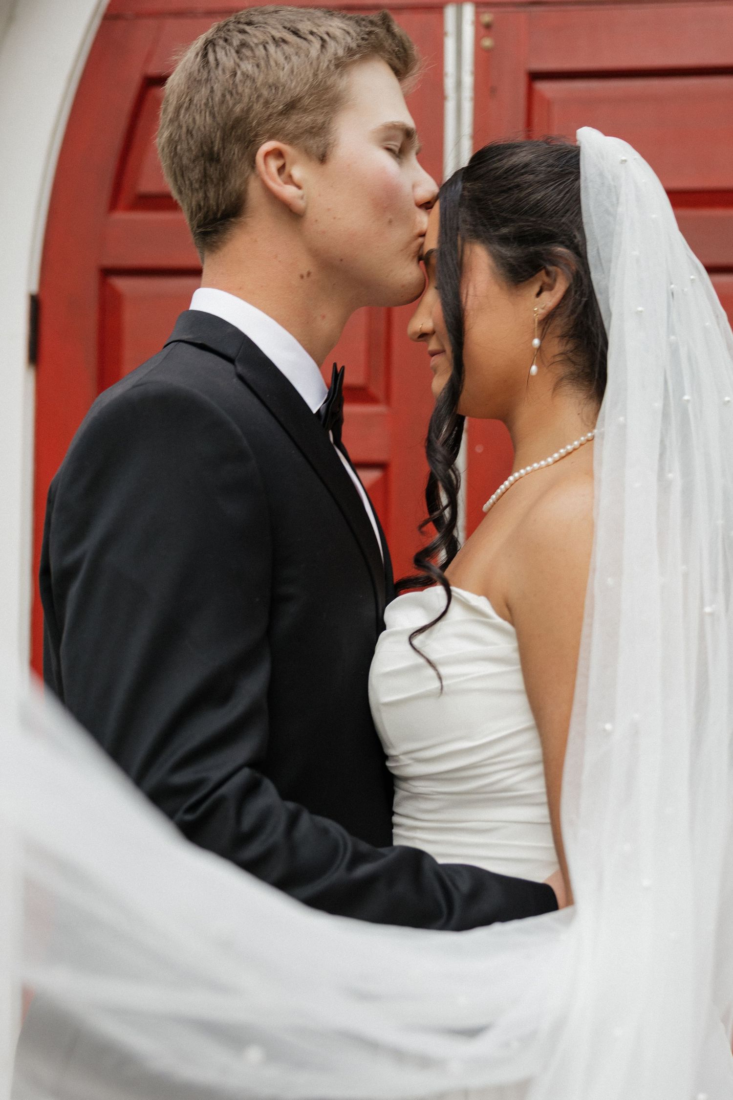 Groom kisses bride on the forehead in front of red doors, bride wearing pearl jewelry and veil.