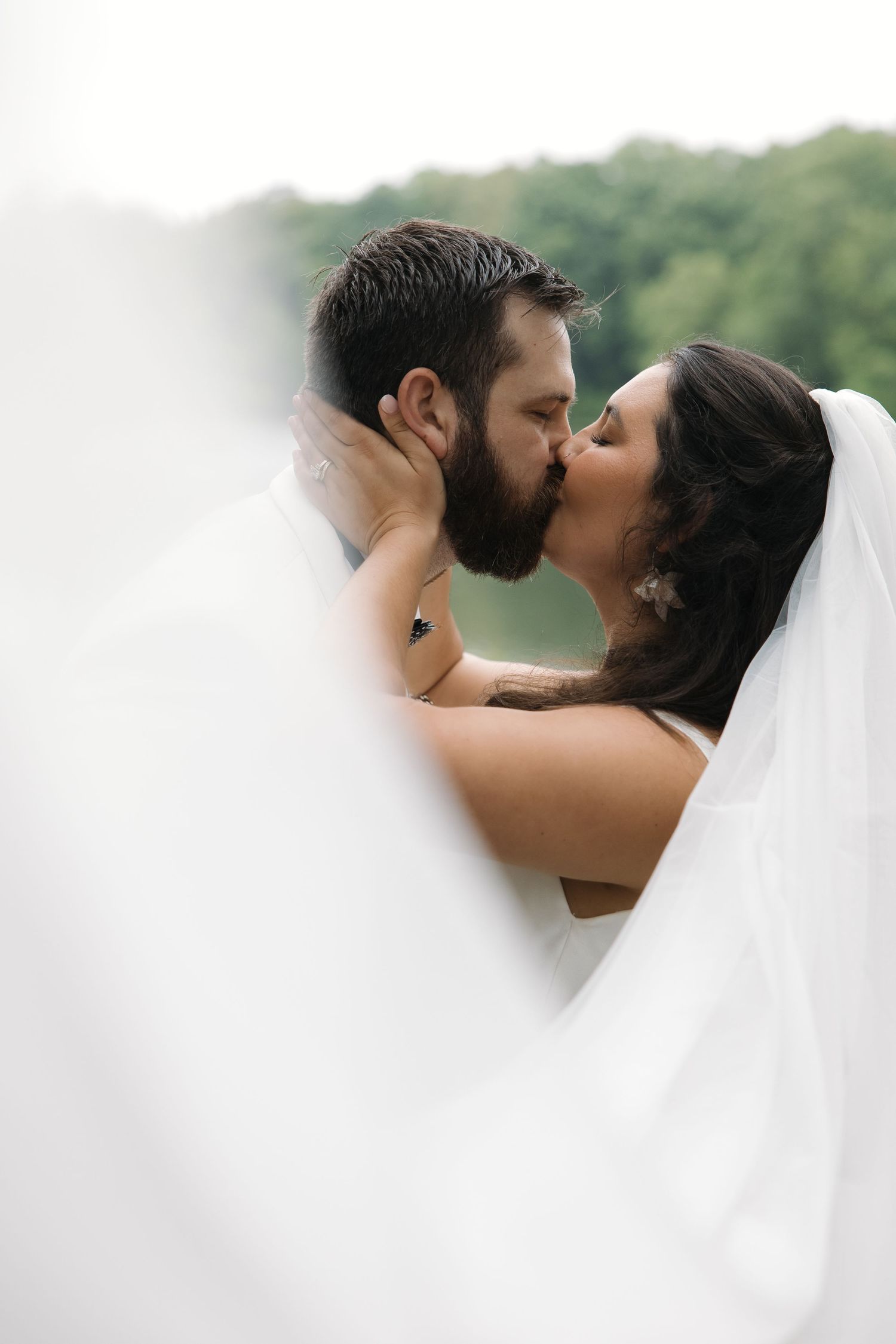 Bride and bearded groom share a passionate kiss outdoors with soft veil framing the moment.