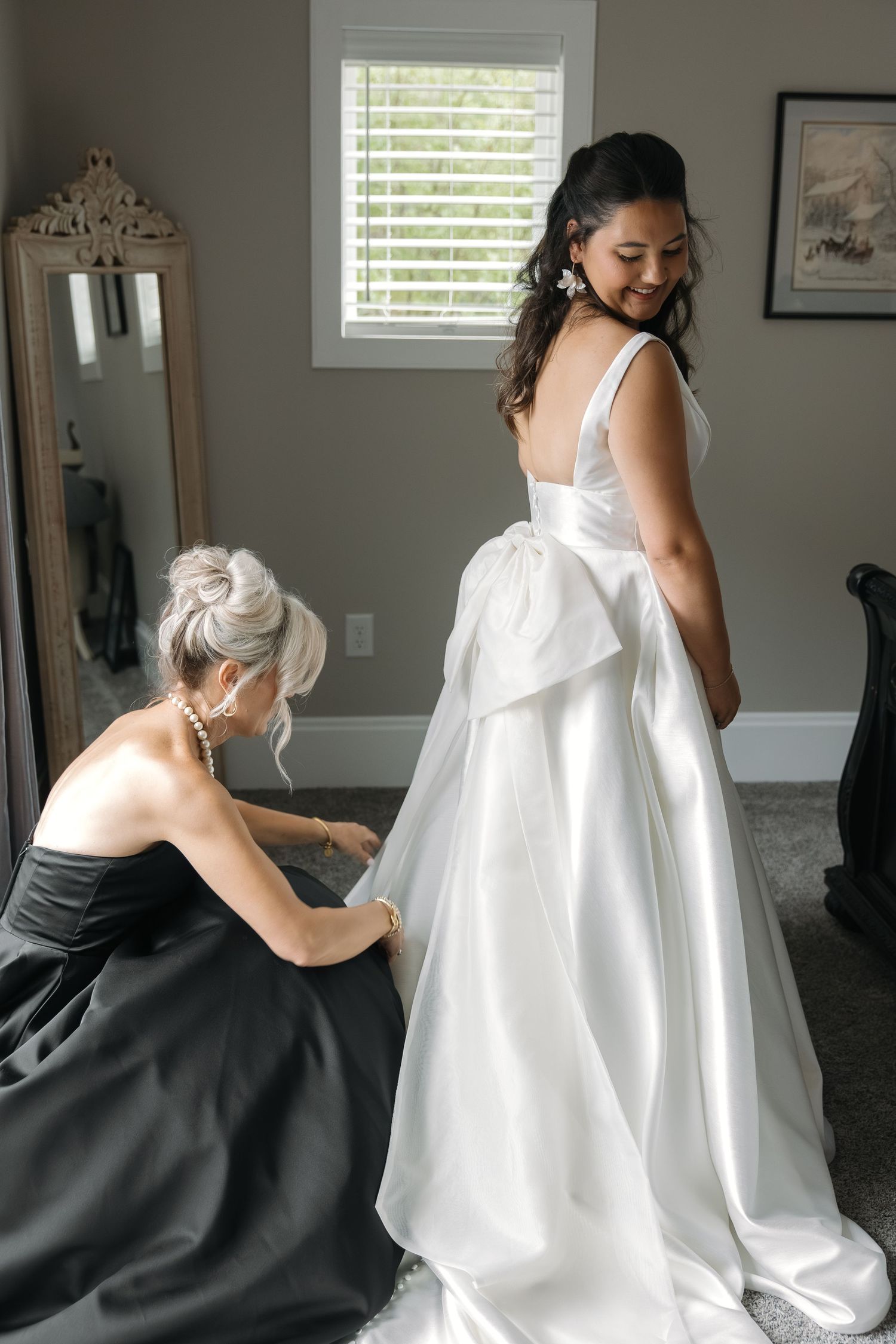 Bridesmaid in black dress fastening the back of the brides elegant white satin wedding gown.