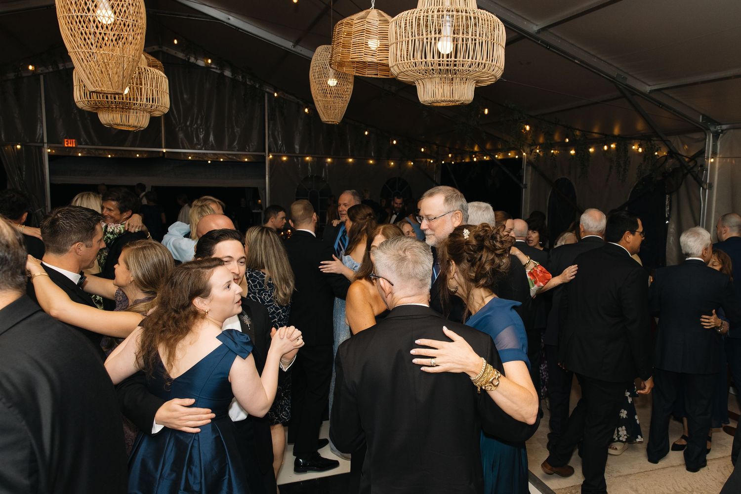 Guests dancing under wicker pendant lights and string lights at elegant tented wedding reception.