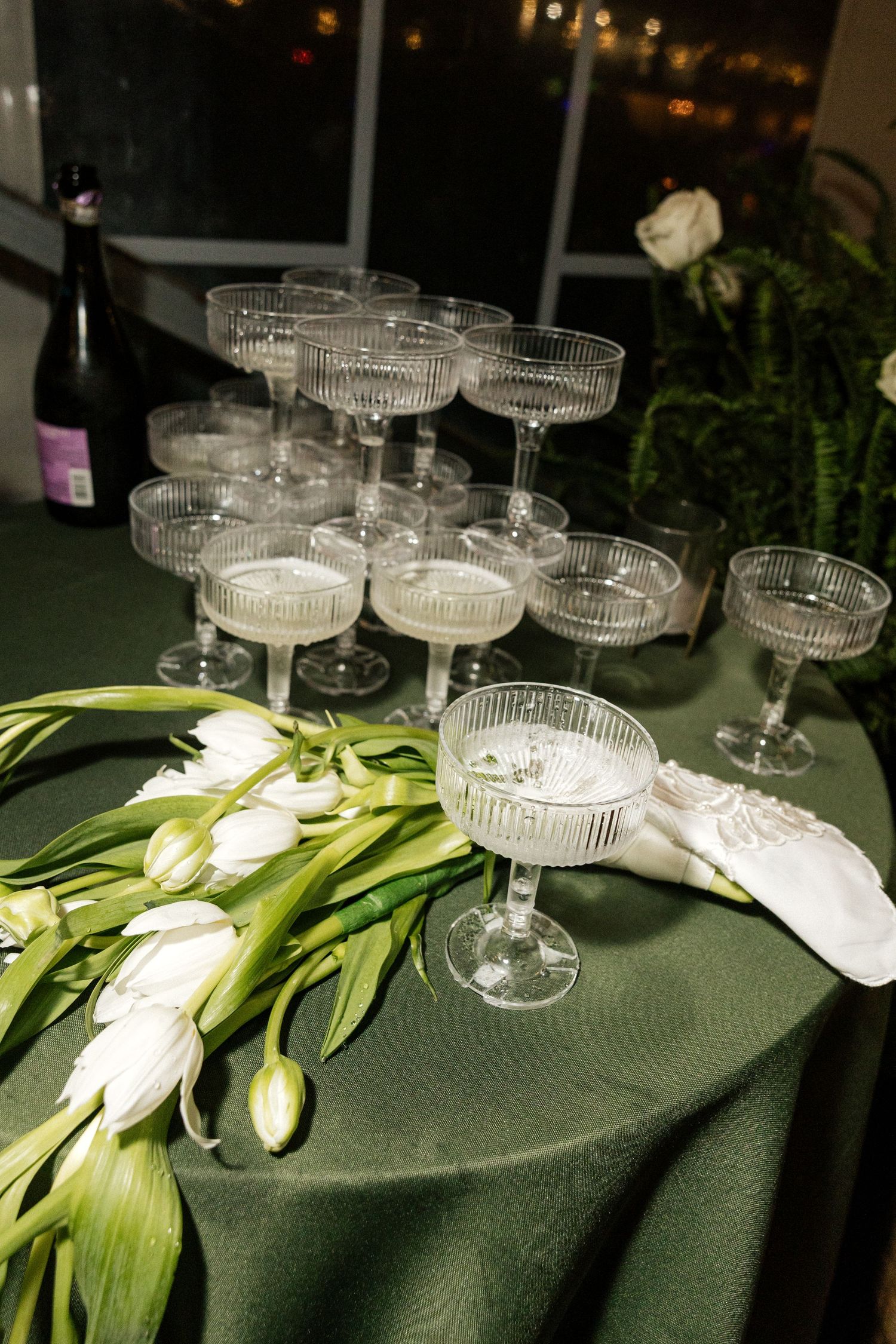 Elegant champagne tower with coupe glasses and white tulips on a green tablecloth at a wedding reception.
