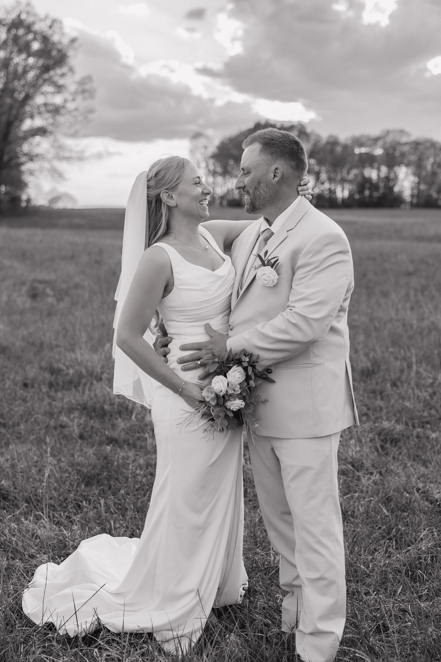 Black and white photo of a smiling bride and groom embracing in an open field at sunset.