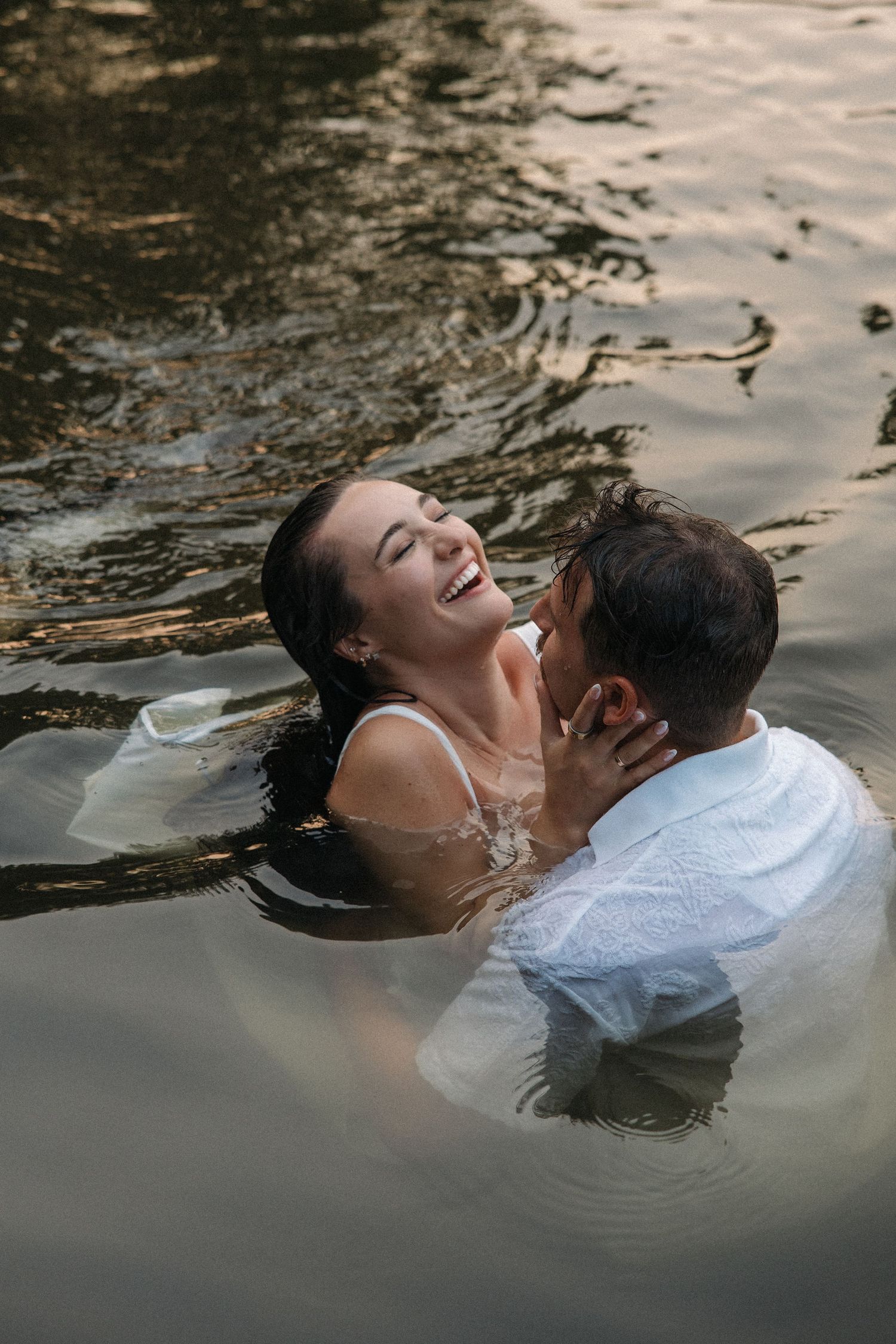 Couple laughing and embracing in water, woman in white dress, man in white shirt, romantic outdoor photo.
