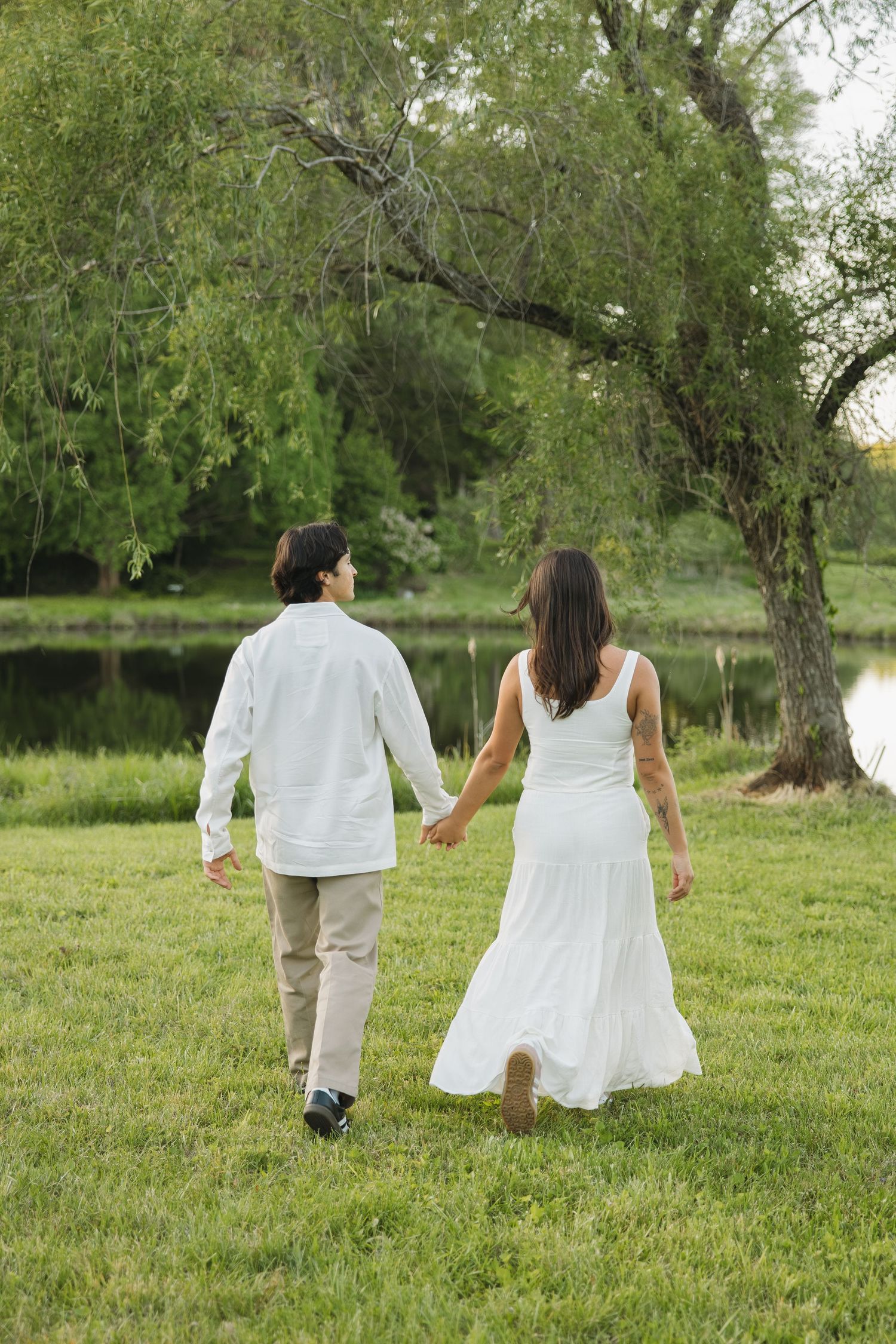 Couple holding hands walking away toward a willow tree by a pond, woman in flowing white dress.