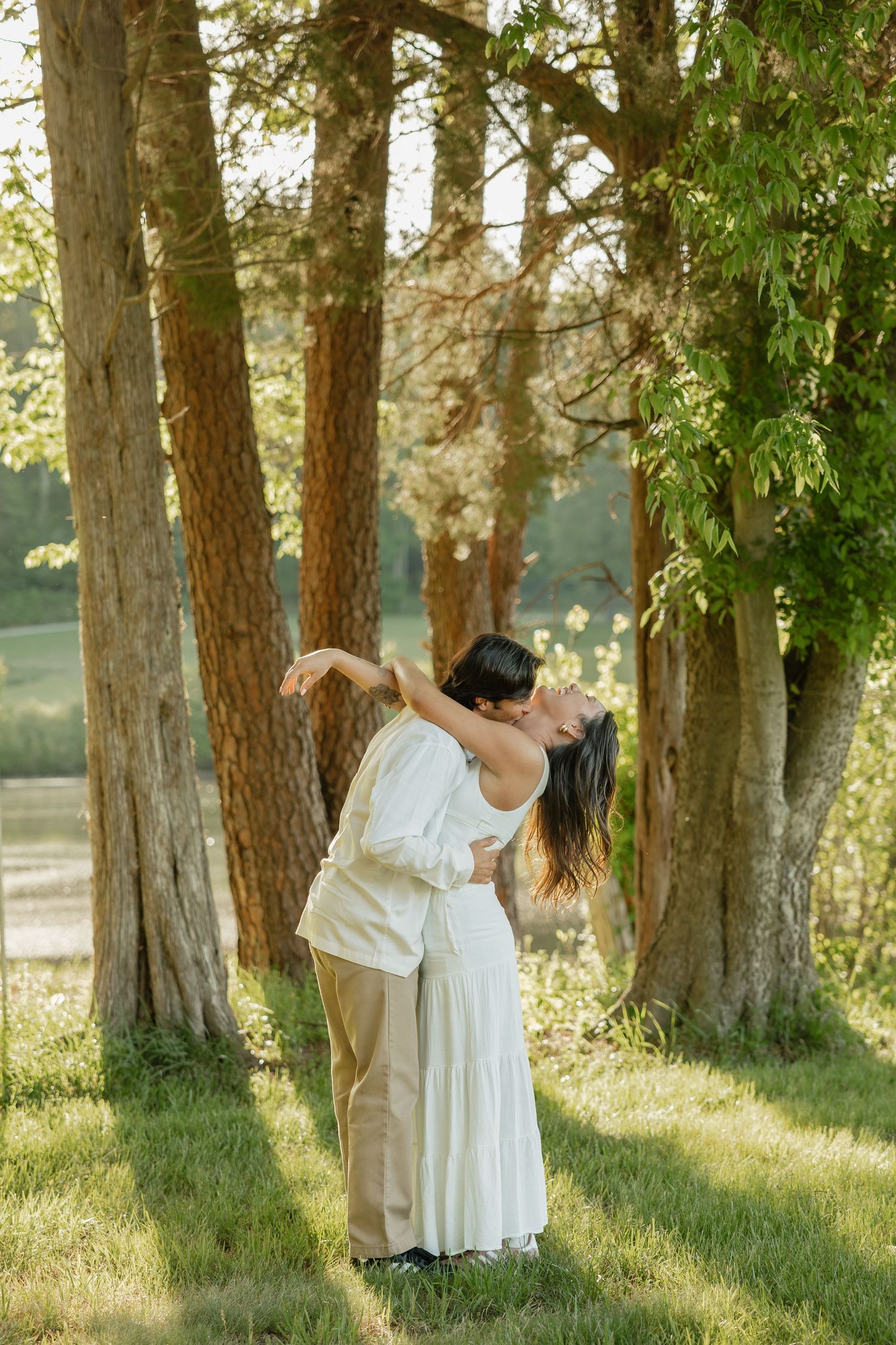 Couple sharing a romantic kiss among tall trees in a sunlit park, woman in white dress leaning back gracefully.