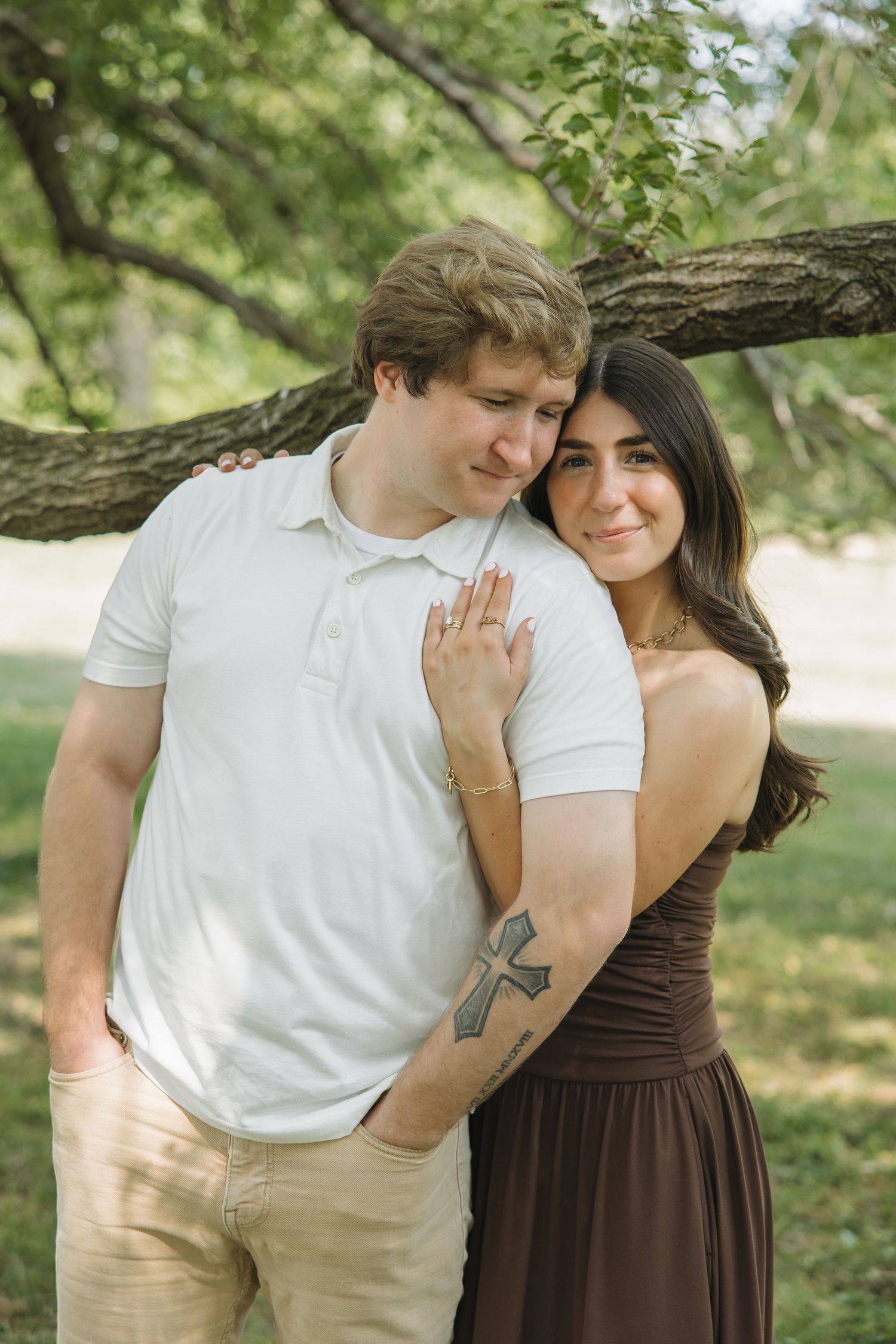 A couple shares a sweet moment under a tree, woman with cross arm tattoo in brown dress smiling warmly.