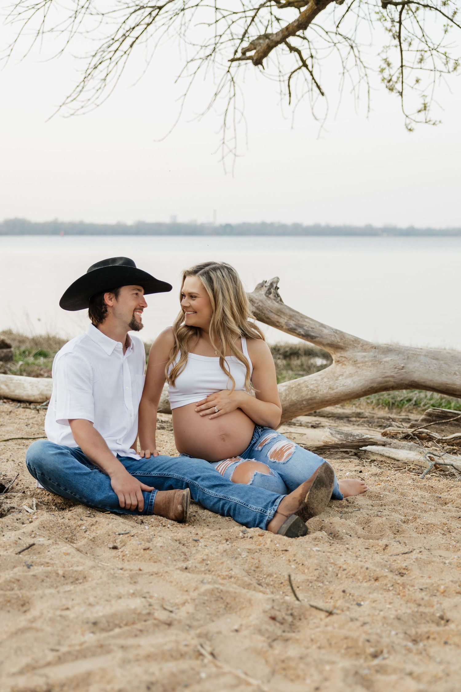 Happy couple sitting on sandy beach by lake, man in cowboy hat gazing at pregnant partner holding her belly.