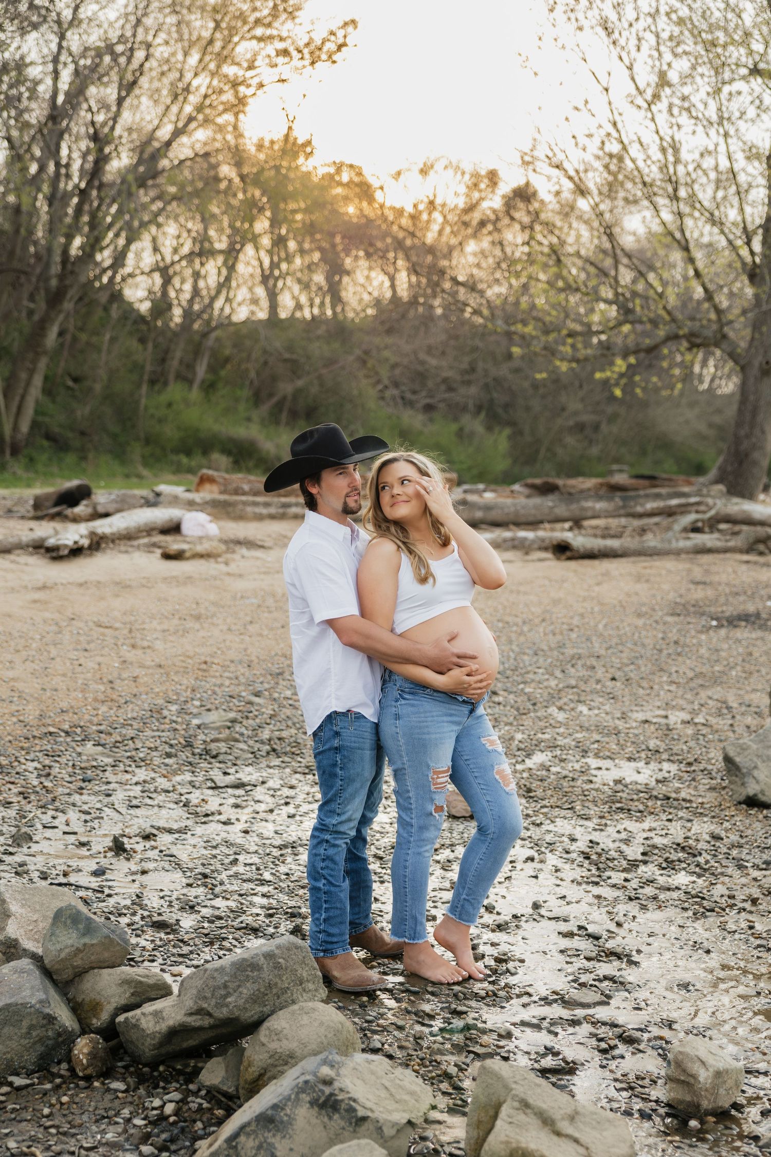 Couple embracing at sunset on rocky beach, pregnant woman looking up at man in black cowboy hat.