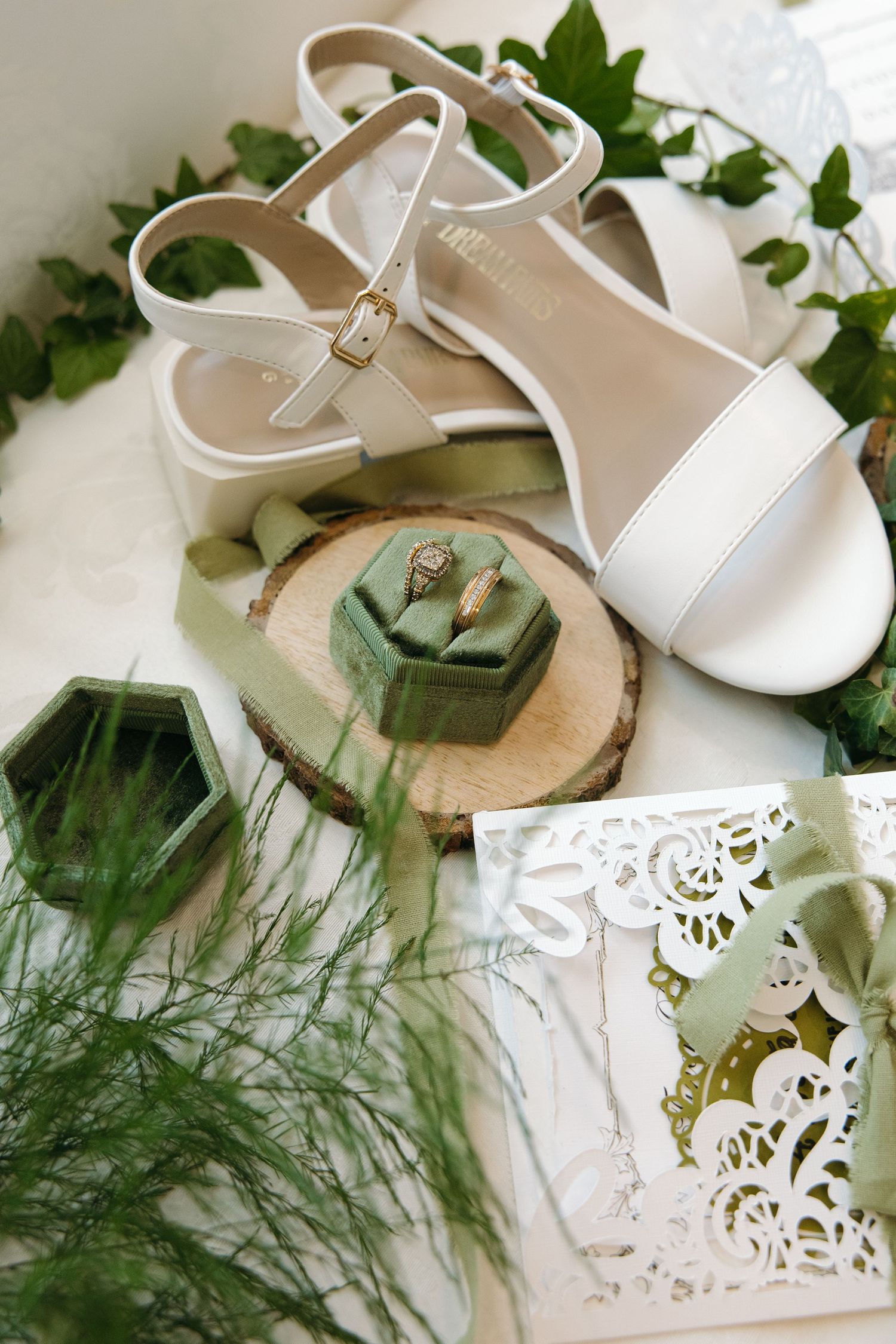 White bridal sandals with gold buckles beside a green velvet ring box with diamond ring on wood slice.