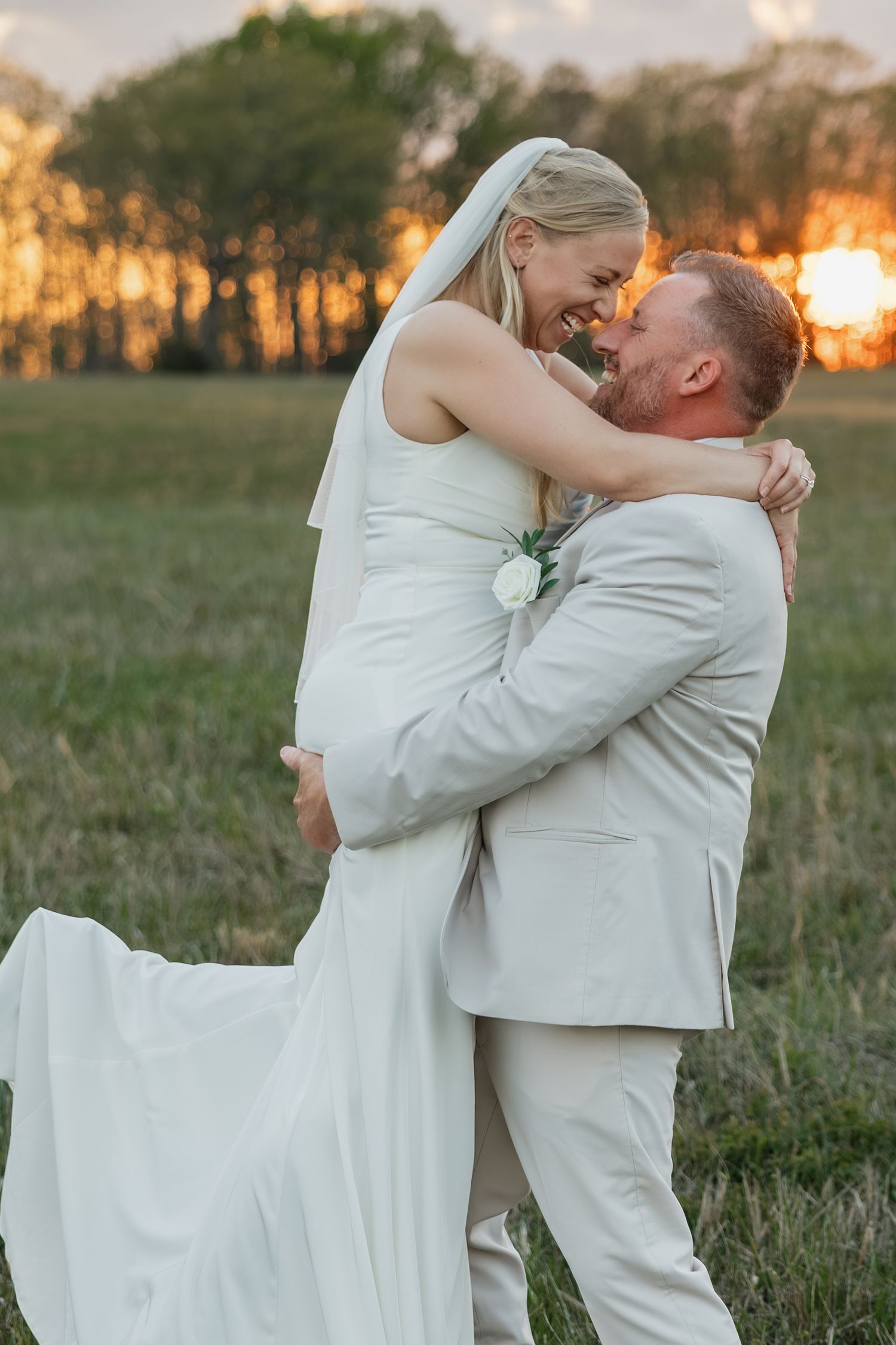 Groom lifting bride in white gown and veil at golden hour sunset in a green field.