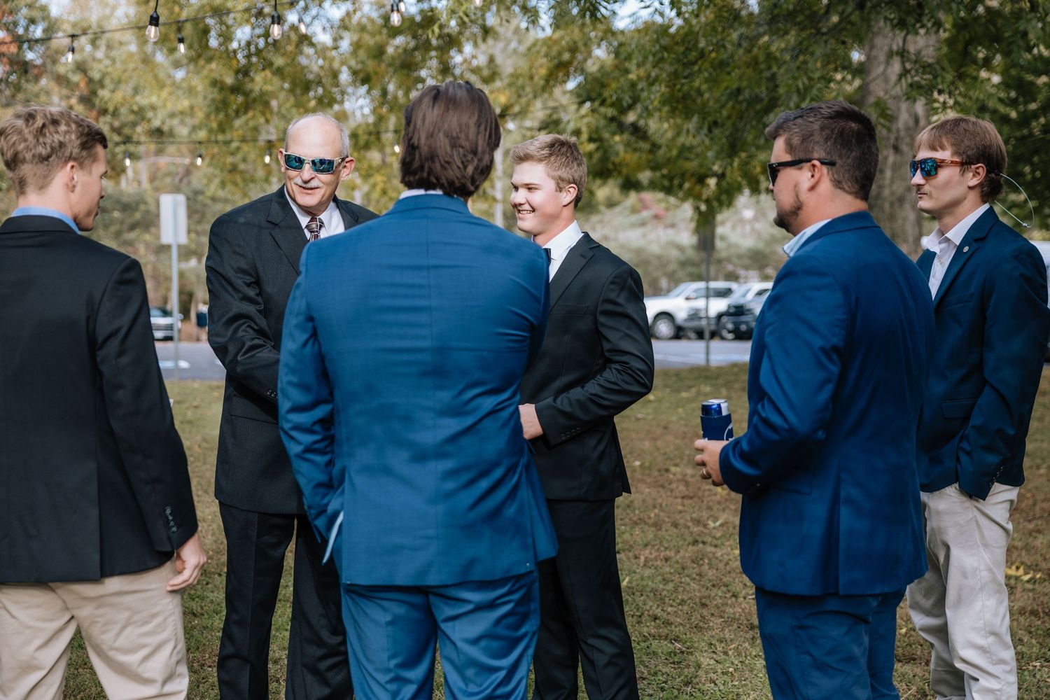 Groomsmen in navy suits and sunglasses gather outdoors chatting before a wedding ceremony.