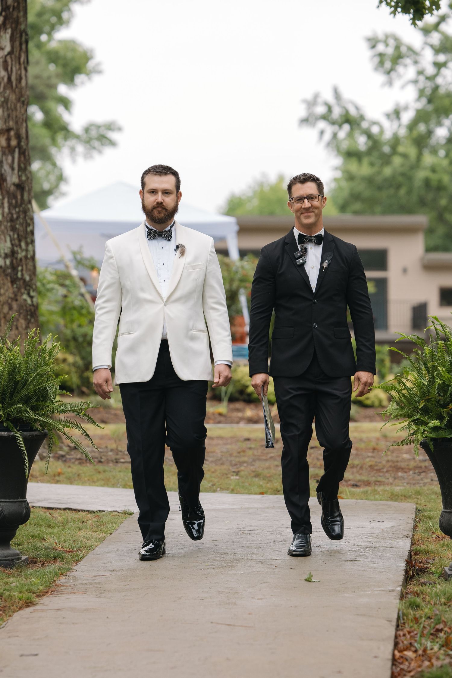 Two grooms walking down the aisle, one in white tuxedo jacket and one in black tuxedo at outdoor wedding.