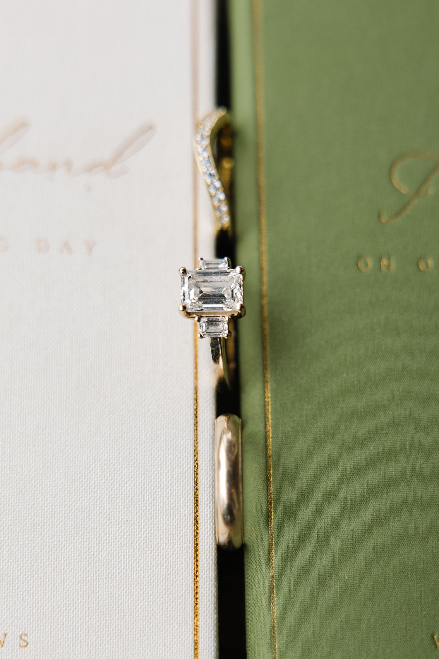 Emerald cut diamond engagement ring with diamond band resting between cream and green wedding vow books.