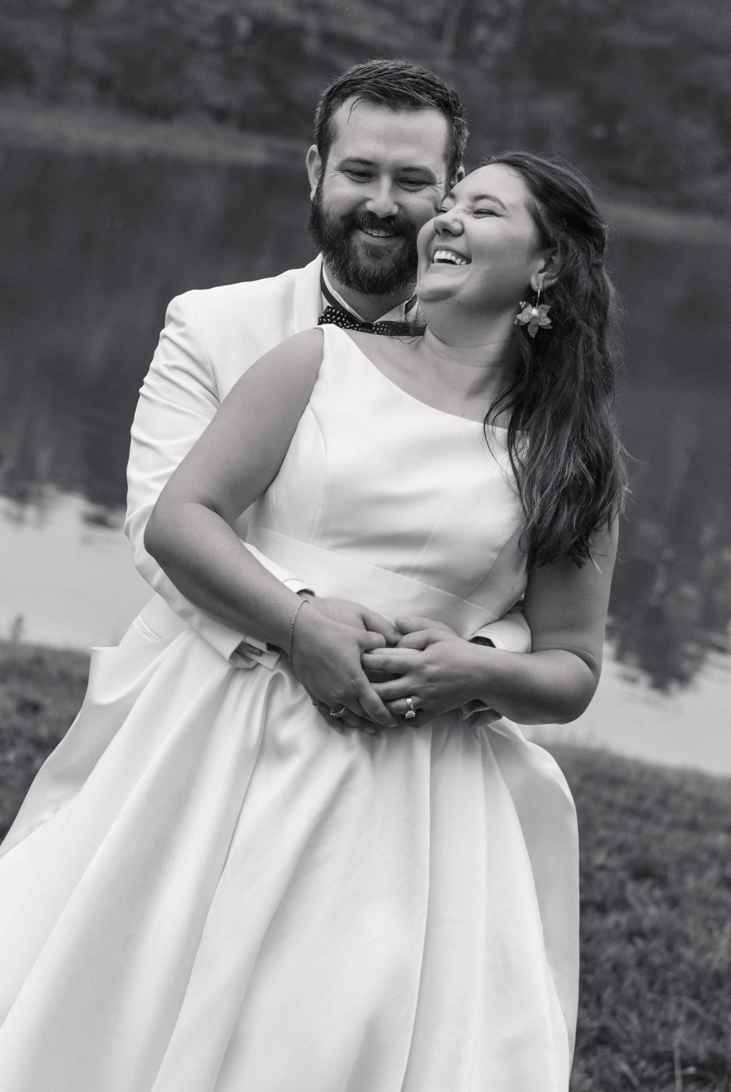 Black and white photo of smiling bride and bearded groom embracing warmly by a lakeside.