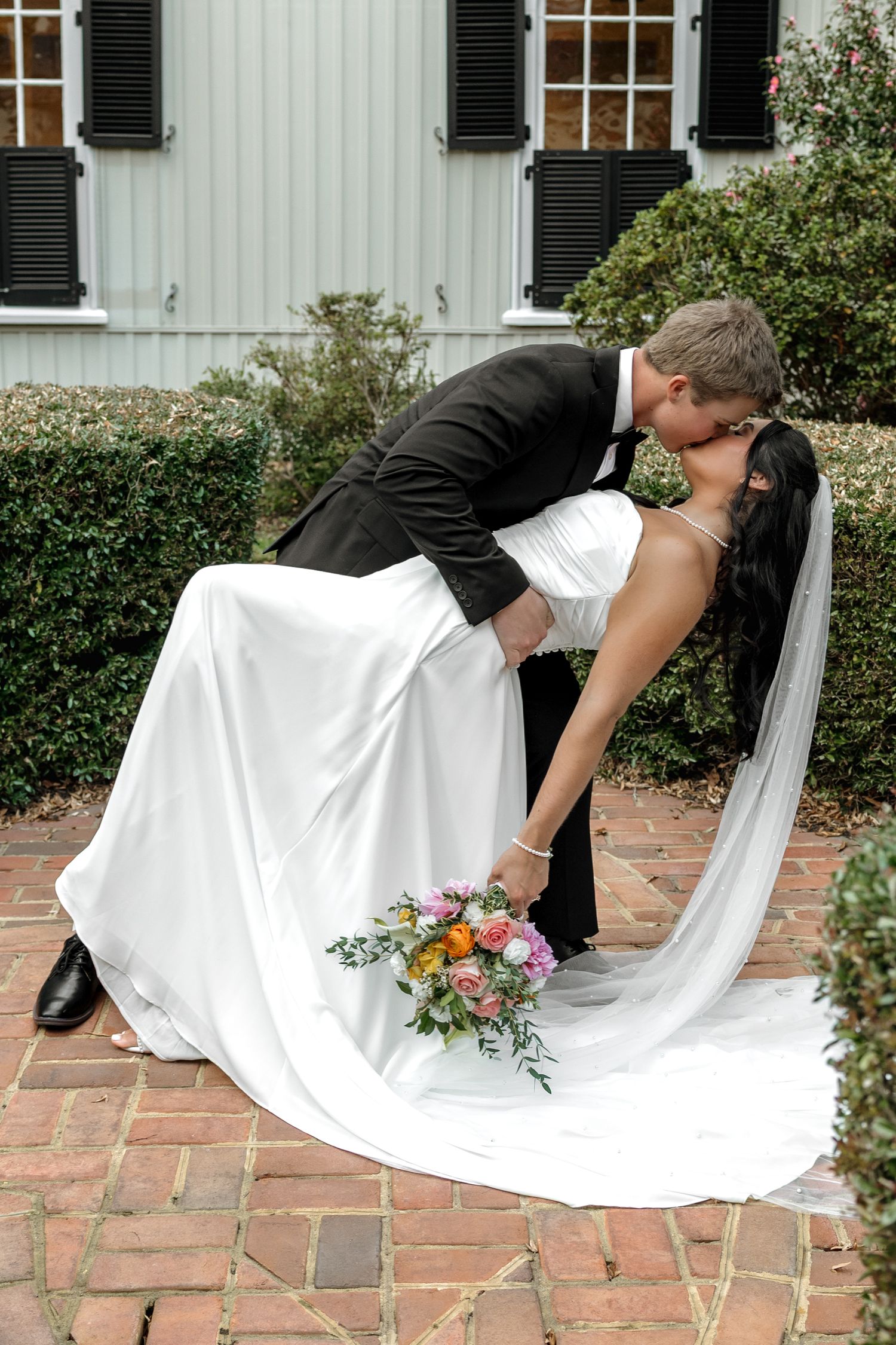 Groom dips bride for a kiss on brick patio outside white building with black shutters, bride holds colorful bouquet.