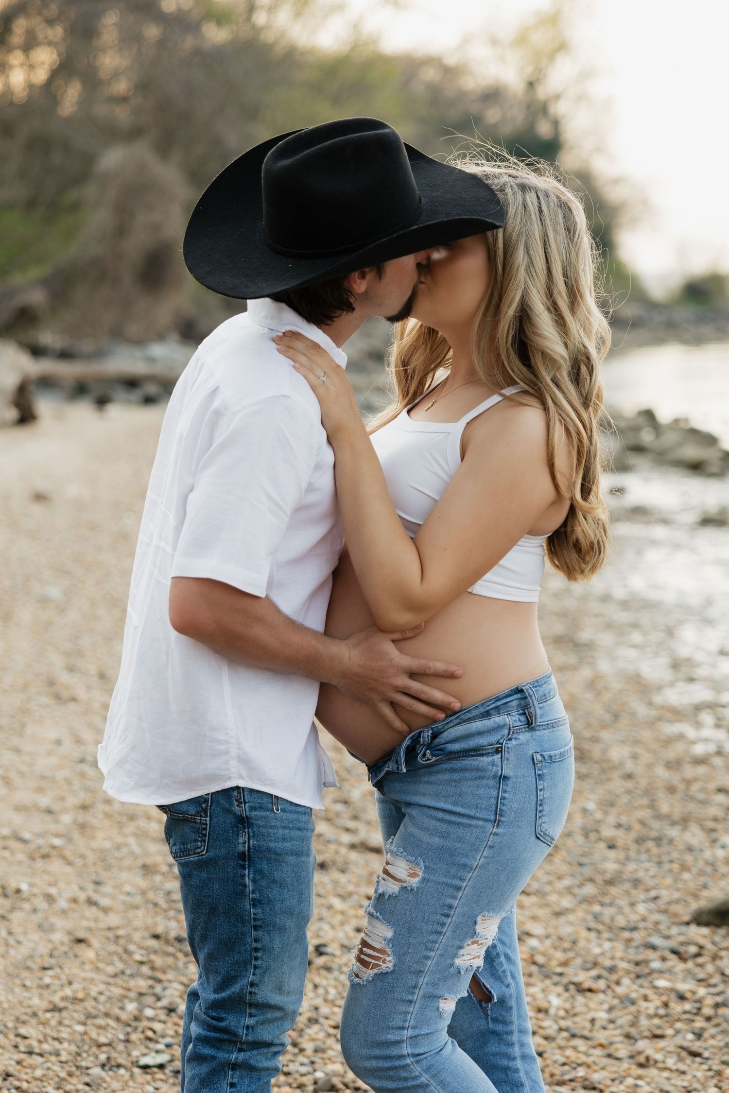 Couple sharing a kiss on rocky beach, man in black cowboy hat embracing pregnant woman's baby bump.