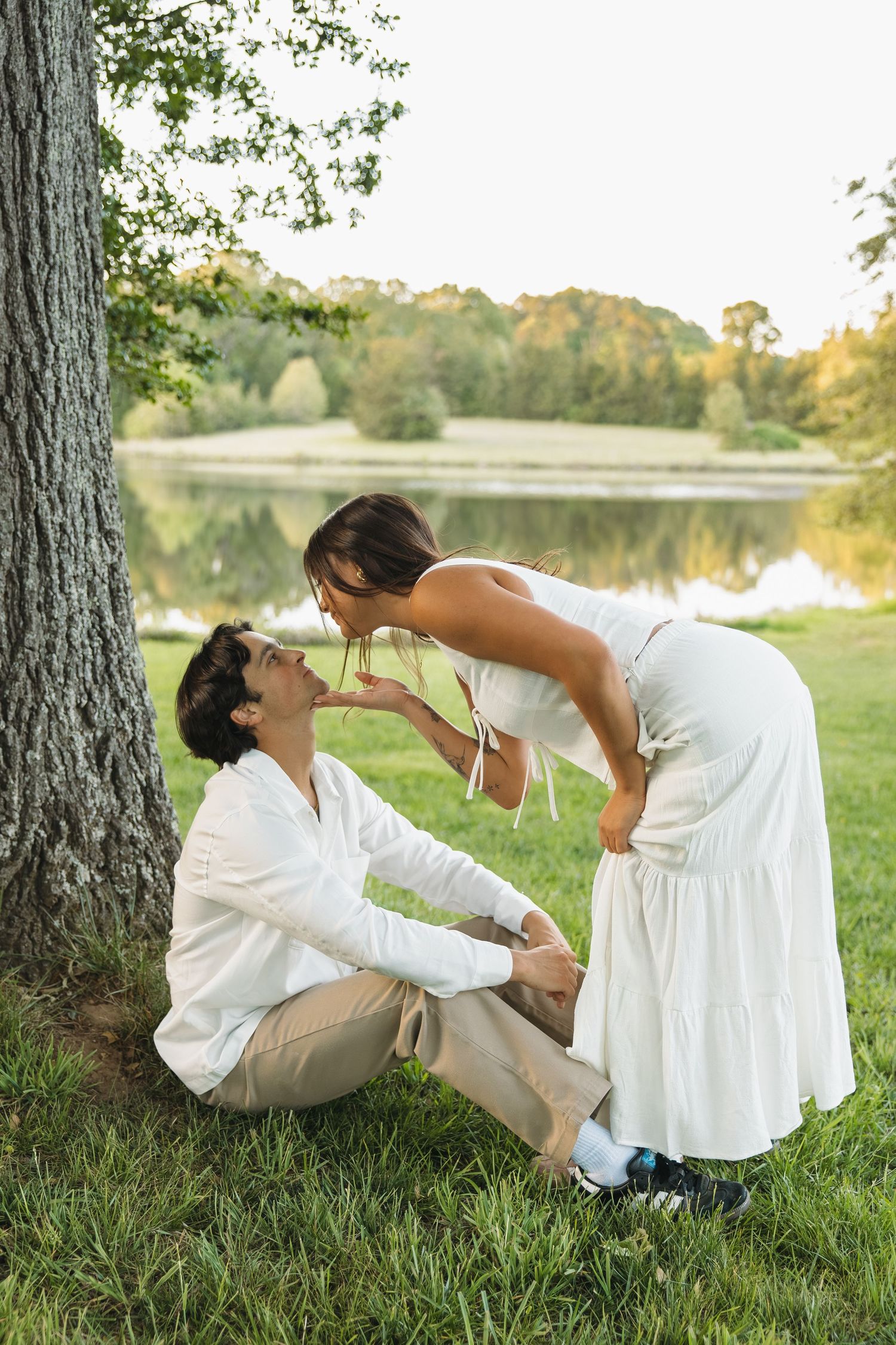 A couple shares a tender moment near a tree by a serene lake during golden hour.