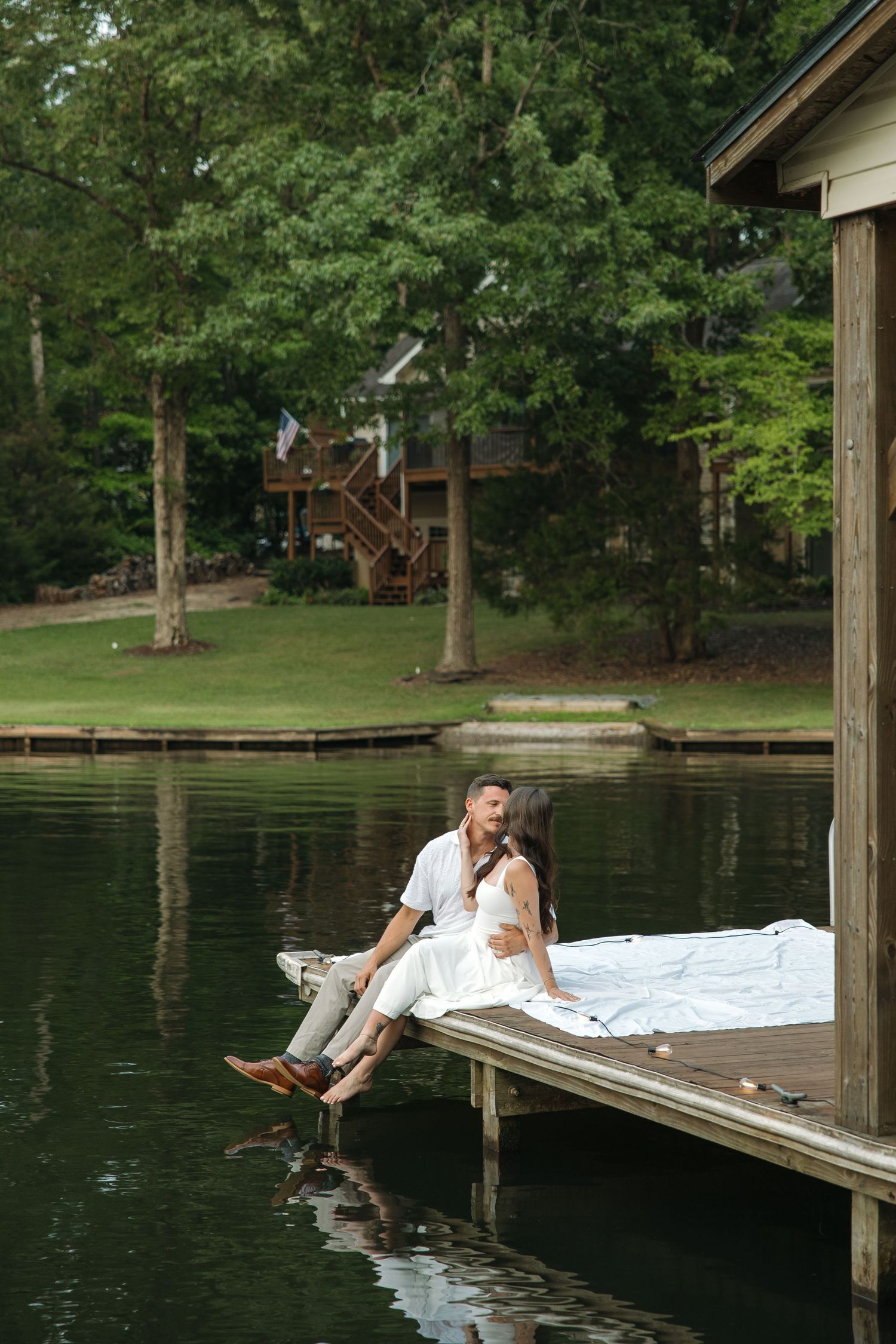Couple sitting on a wooden dock by a calm lake, surrounded by lush green trees and a lakeside cabin.