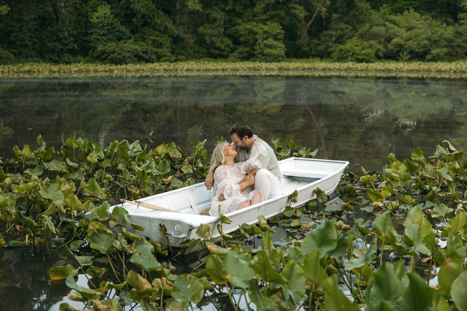 A couple kisses intimately in a white rowboat nestled among lily pads on a peaceful lake with forest backdrop.