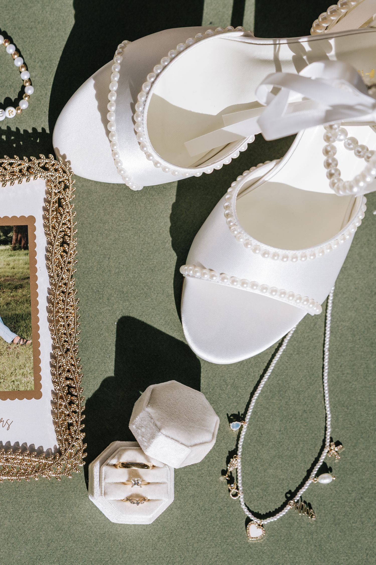 Bridal flat lay featuring pearl-adorned heels, gold ring box, jewelry, and wedding accessories on green.