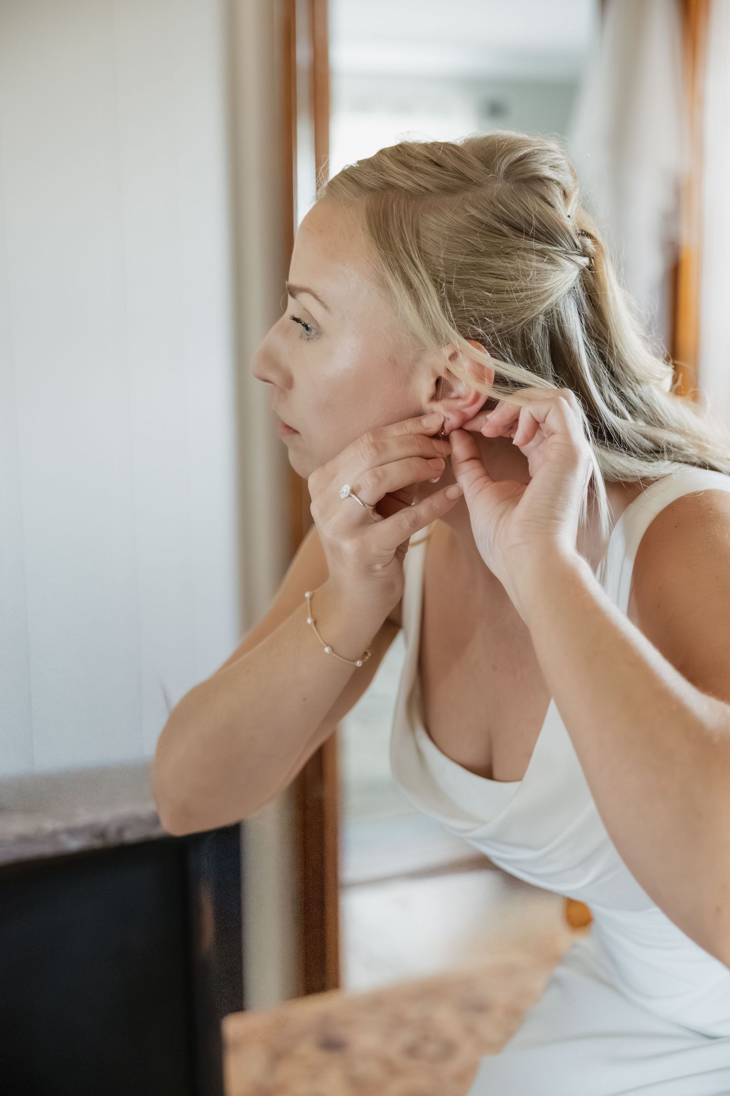 Elegant blonde bride putting on earrings while looking in mirror during wedding day preparations.